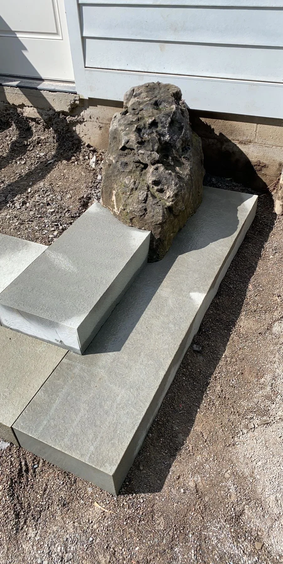 A large irregularly shaped rock placed on a concrete walkway, with concrete blocks surrounding it, in front of a house with white siding.