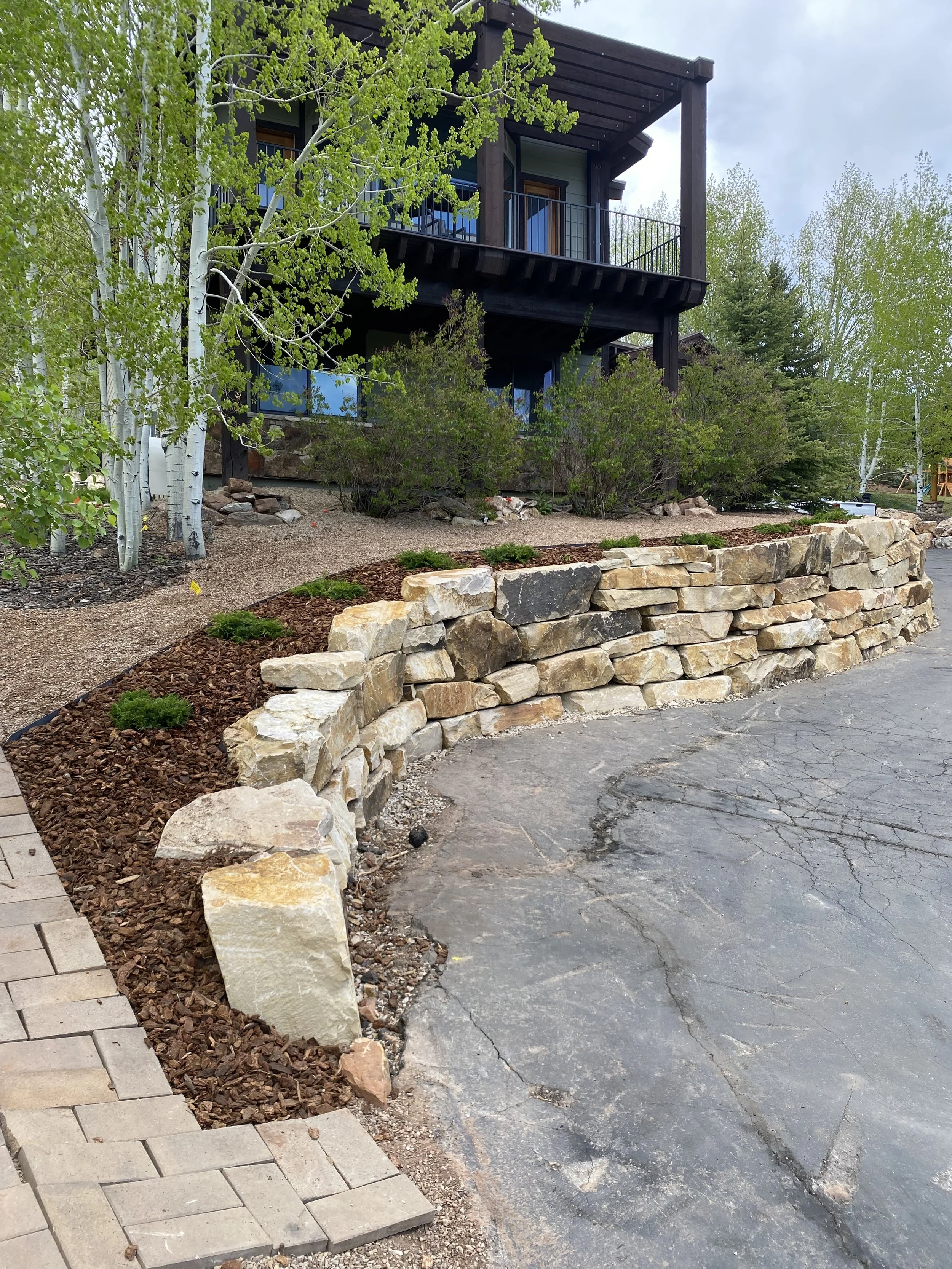 A stone retaining wall in front of a house with a dark wooden exterior and a balcony, surrounded by trees and landscaping.