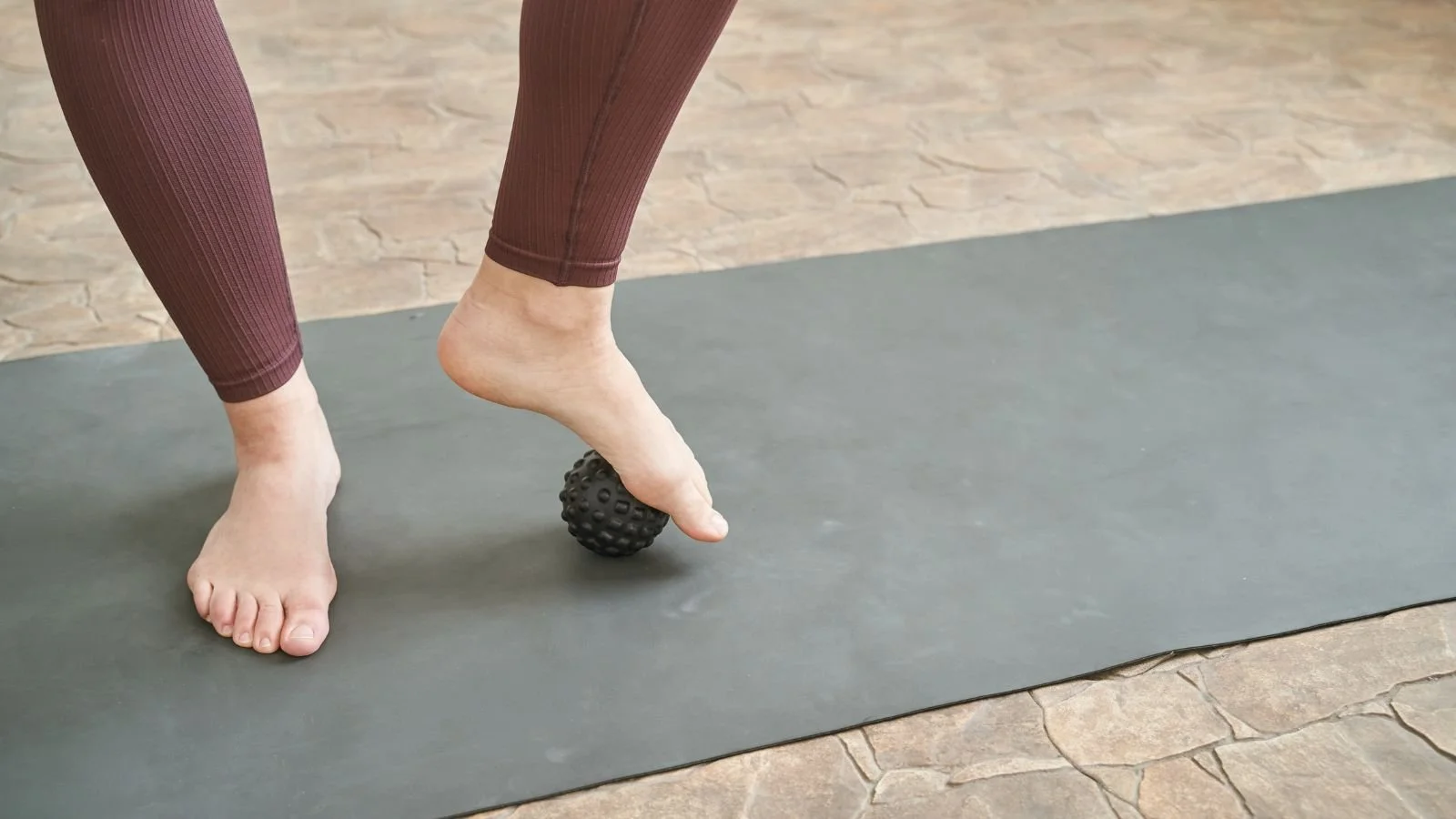 Person using a black massage ball on the arch of their foot during a yoga or stretching session on a gray mat.