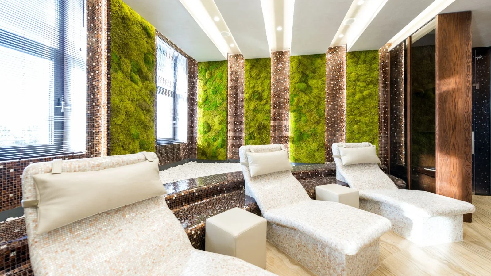 Spa relaxation lounge with three white lounge chairs, small white tables, green moss wall decor, large windows with blinds, and wooden accents.