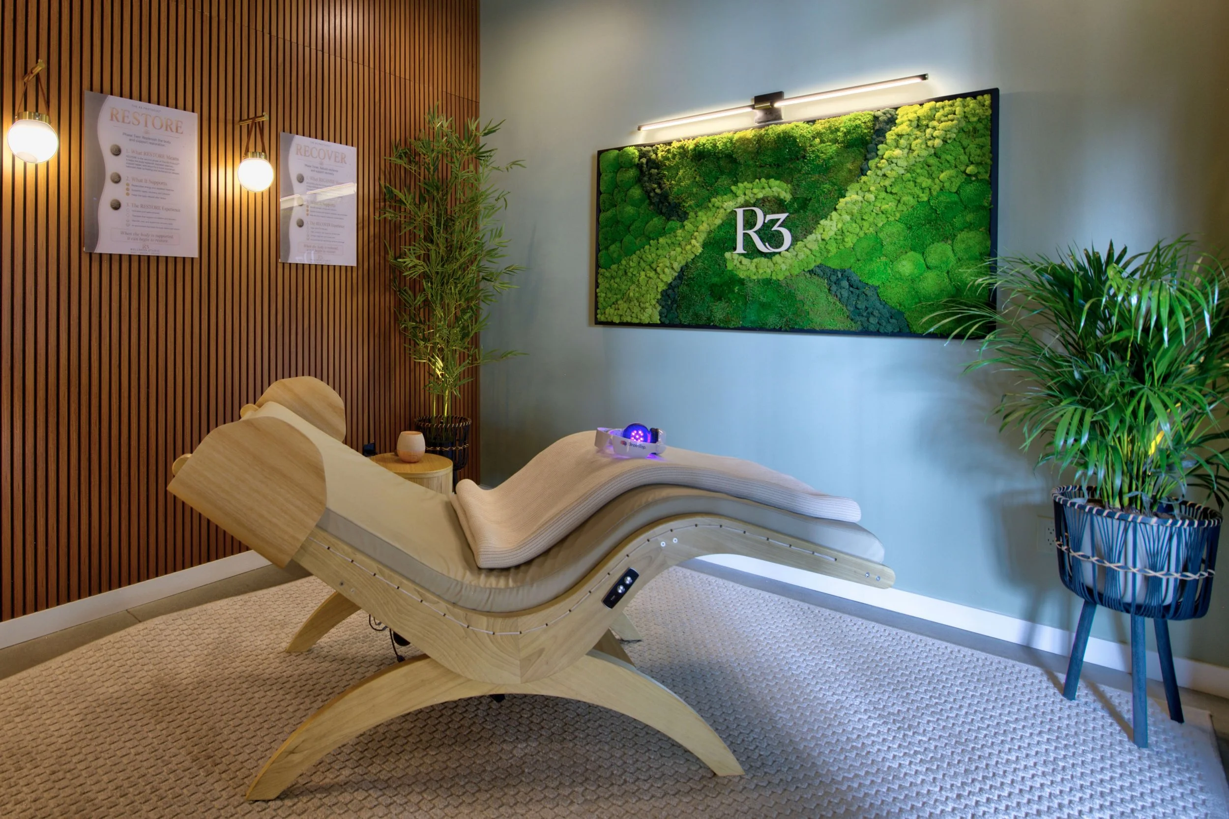 Spa relaxation lounge with three white lounge chairs, small white tables, green moss wall decor, large windows with blinds, and wooden accents.