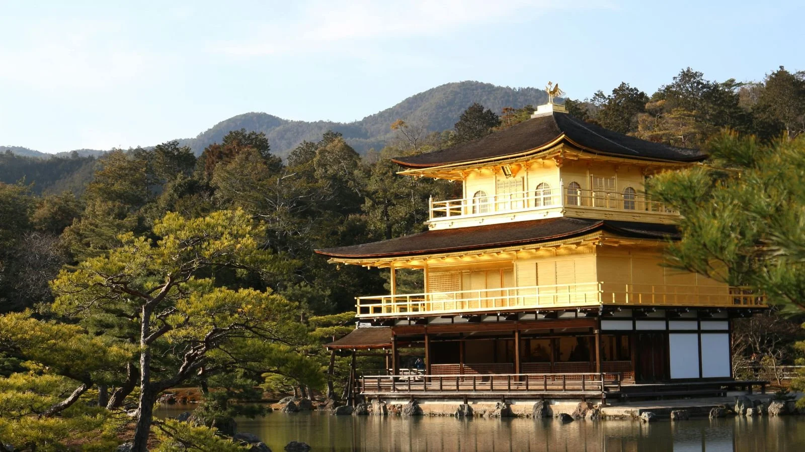 Golden temple with traditional Japanese architecture surrounded by trees and water, set against a mountain background in Kyoto, Japan.
