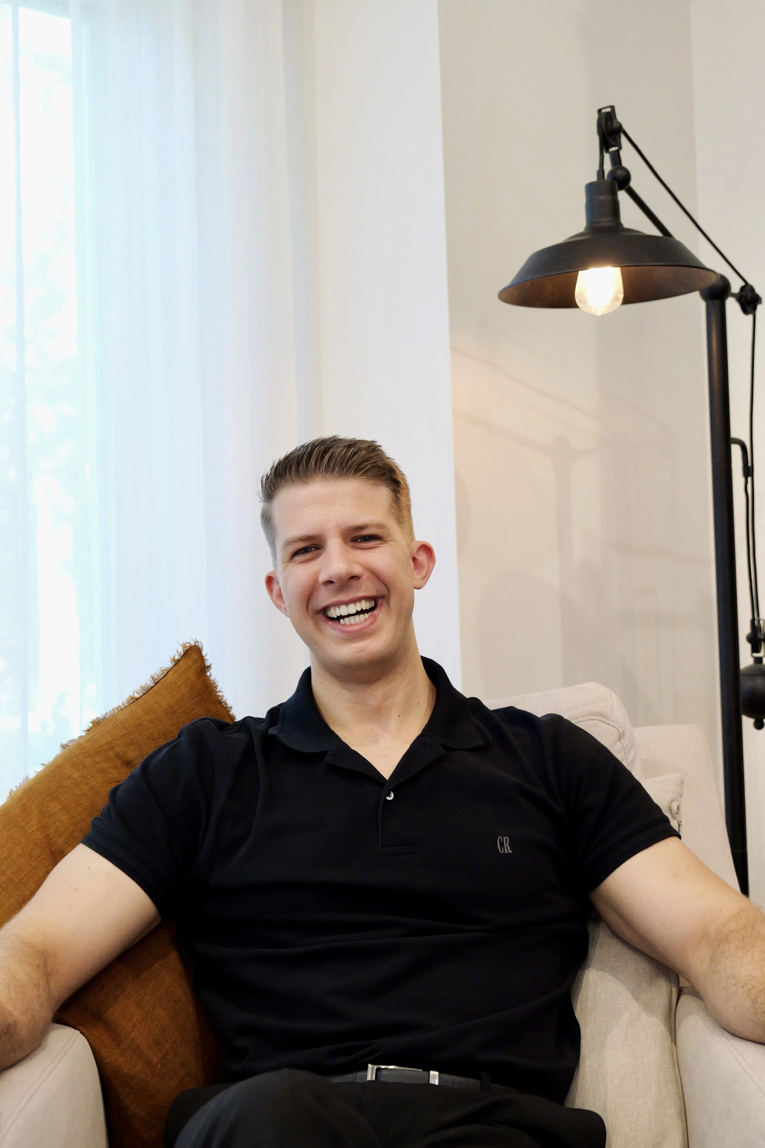 A young man with short light brown hair sitting on a beige couch, smiling broadly at the camera. He's wearing a black polo shirt and is surrounded by cushions, with natural light coming from a window with white curtains behind him. A black floor lamp with an exposed bulb is visible to his right.