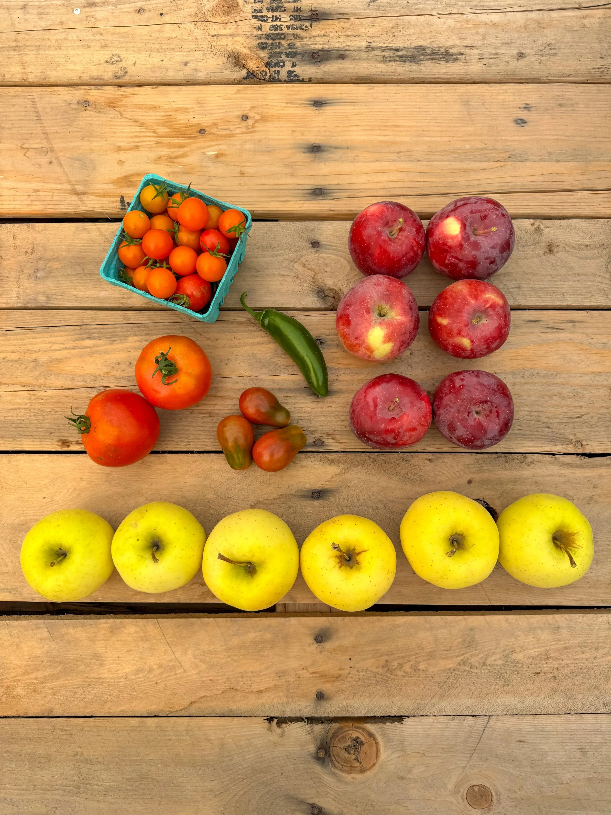 Assorted fresh fruits displayed on a wooden surface, including cherry tomatoes in a blue container, larger round tomatoes, green chili pepper, red apples, and yellow apples.