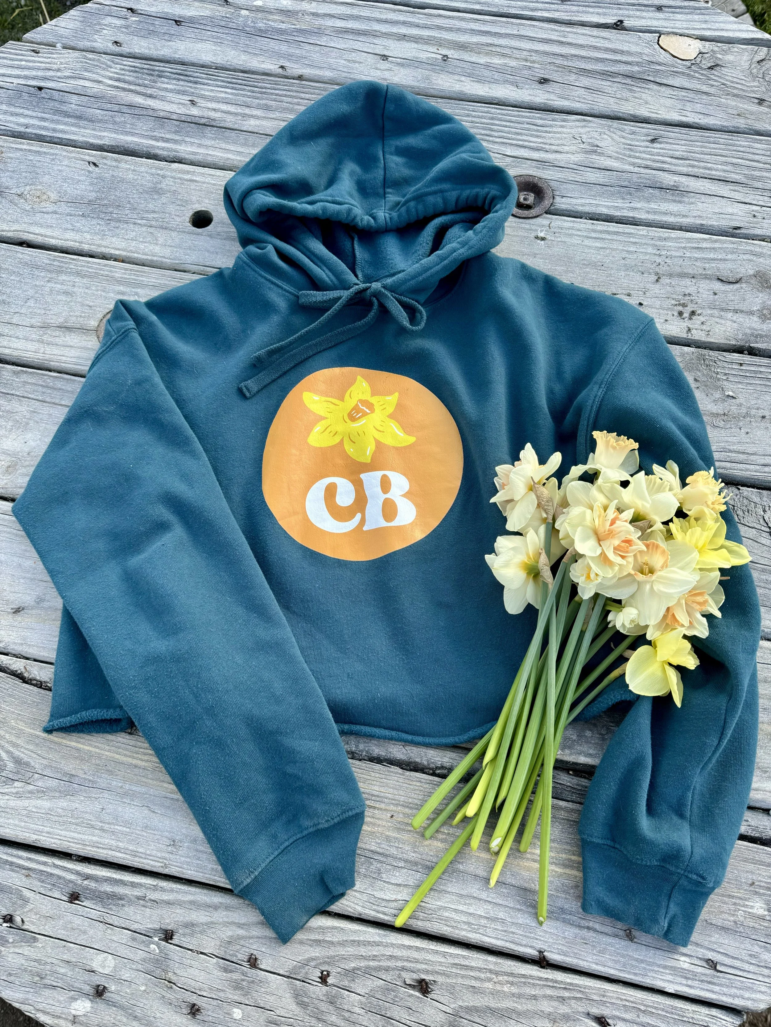 A teal hoodie with a graphic of a yellow flower and the letters 'CB' in white inside an orange circle, placed next to a bouquet of yellow and white flowers on a wooden surface.