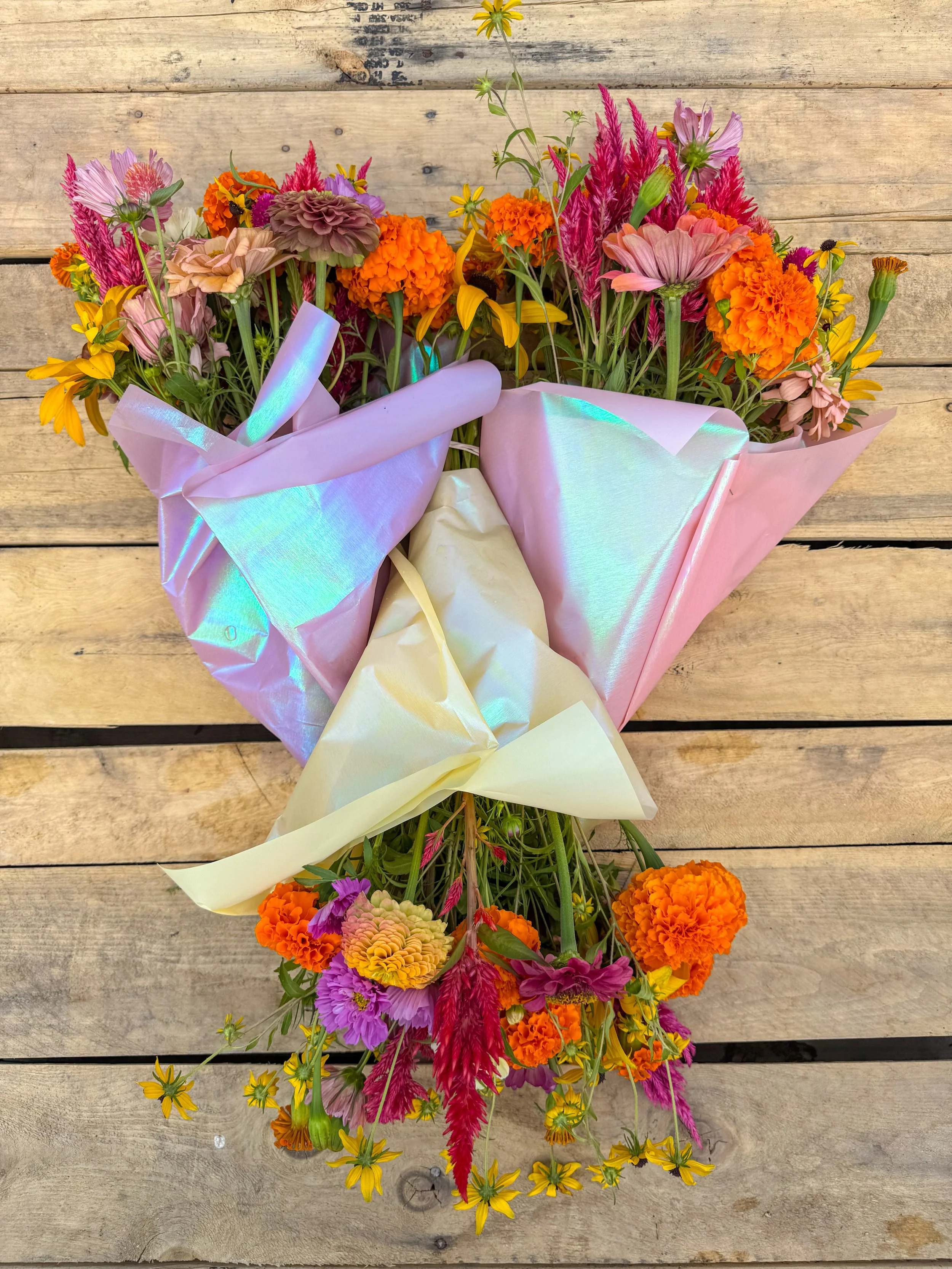Colorful mixed flower bouquet with pink, orange, purple, and yellow flowers wrapped in iridescent and pastel paper, laid on a wooden surface.