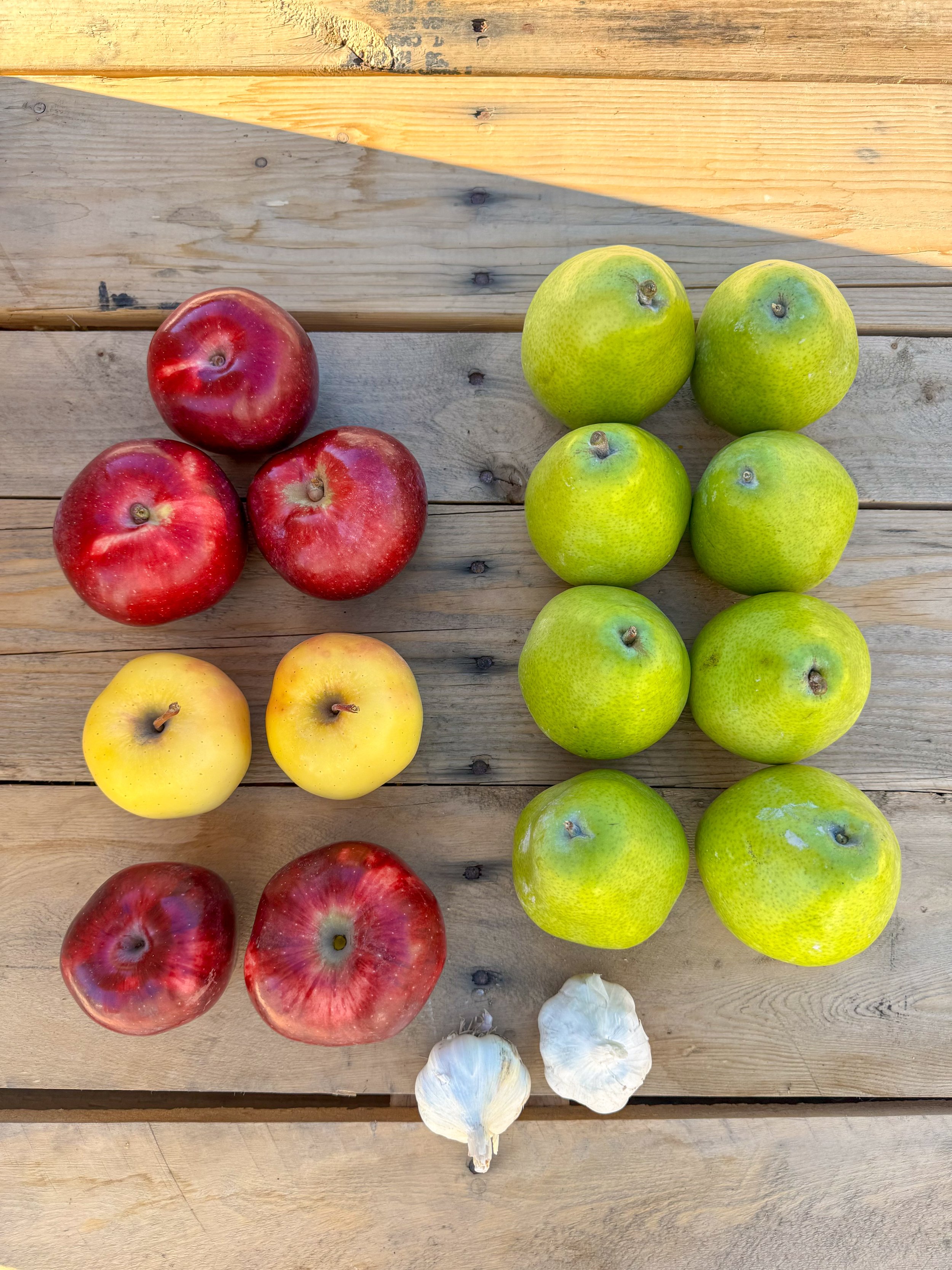 A top-down view of three groups of apples and two garlic bulbs on a wooden surface. The apples are red, yellow, and green.