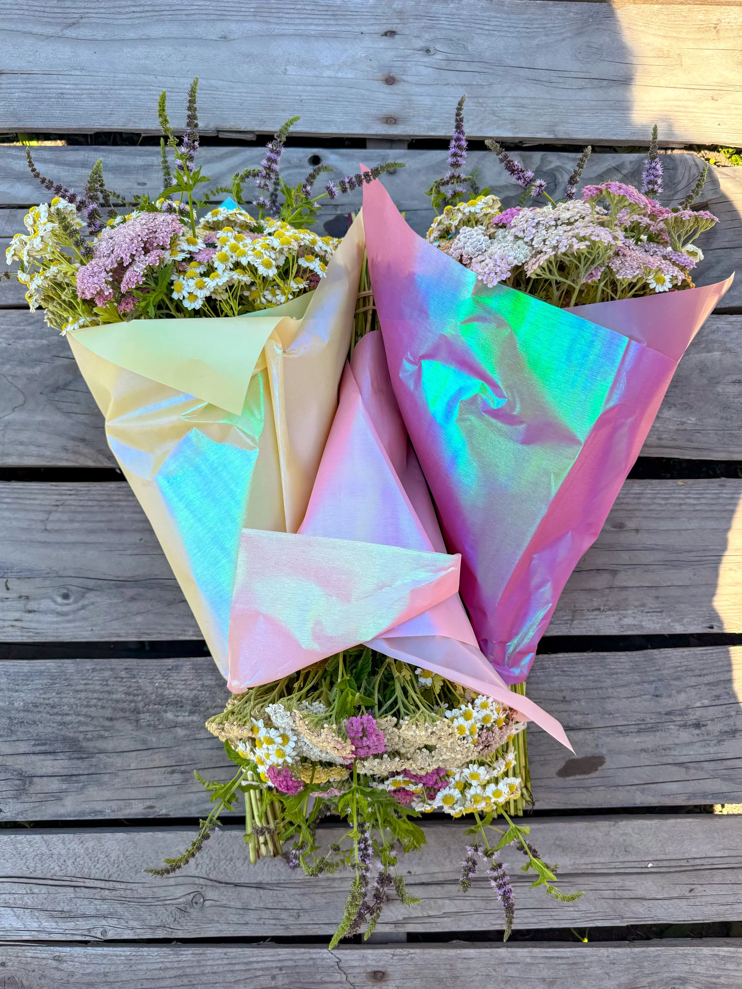 Bouquet of mixed wildflowers wrapped in iridescent pastel-colored paper, placed on a wooden surface.