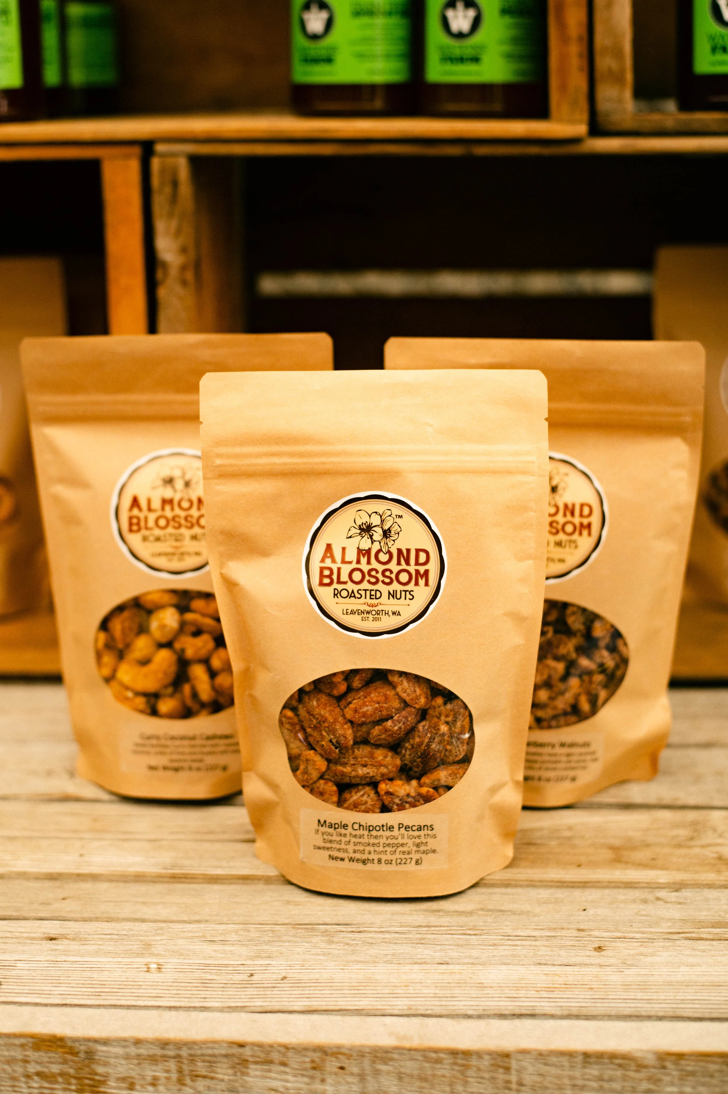 Three bags of roasted almonds on a wooden surface, labeled 'Almond Blossom Roasted Nuts,' with the front bag featuring a label for Maple Chipotle Pecans.