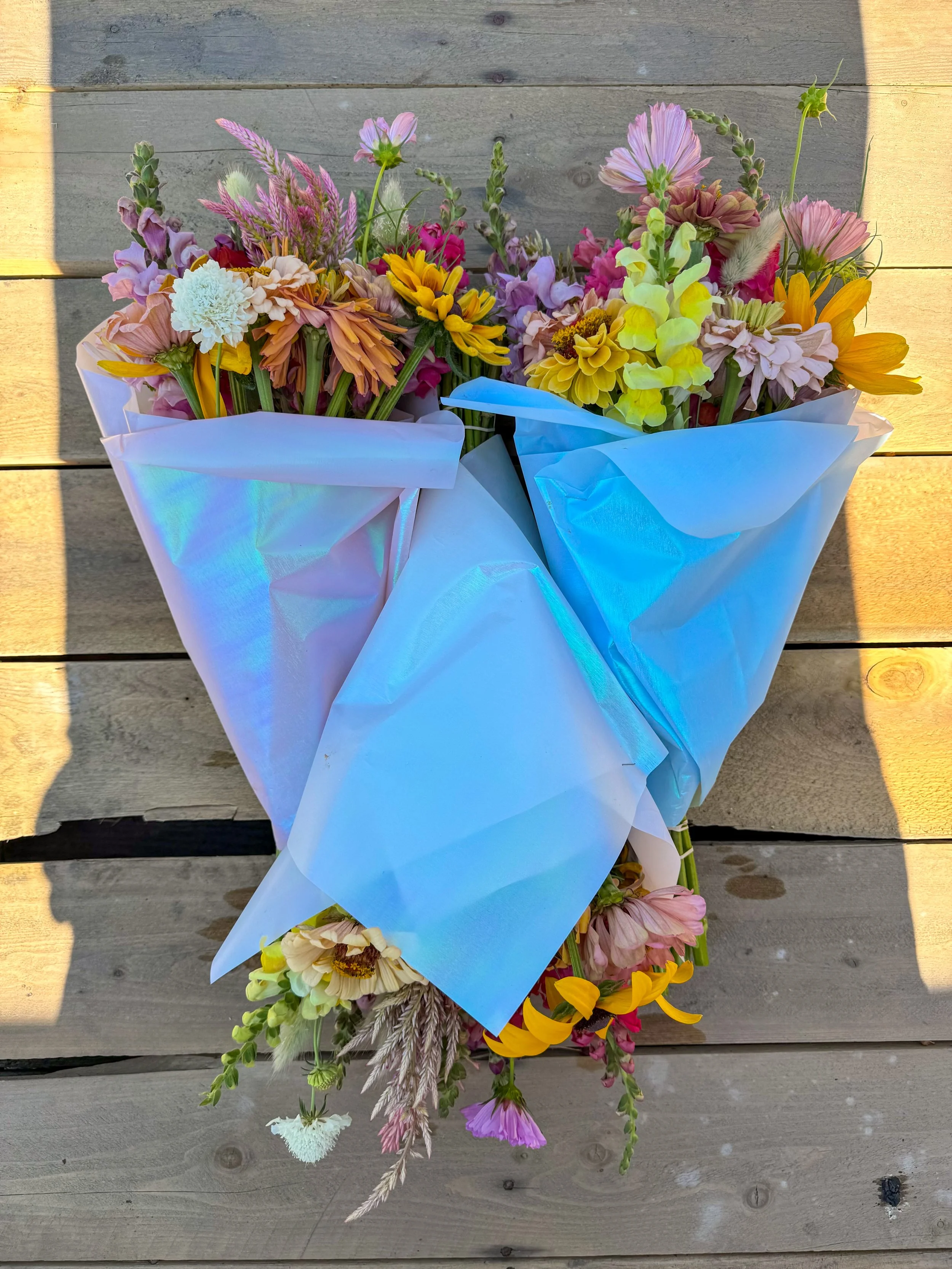 Two bouquets of colorful mixed flowers wrapped in shiny white and blue wrapping paper on a wooden surface.