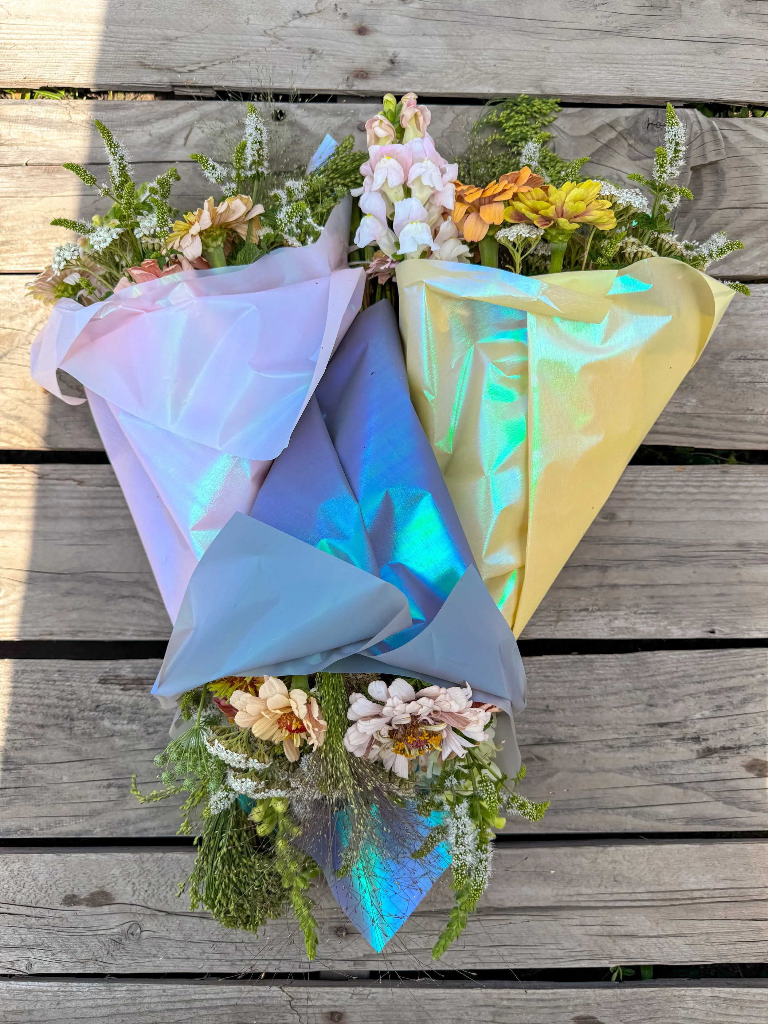 Bouquet of mixed flowers wrapped in iridescent pastel-colored paper on wooden surface.