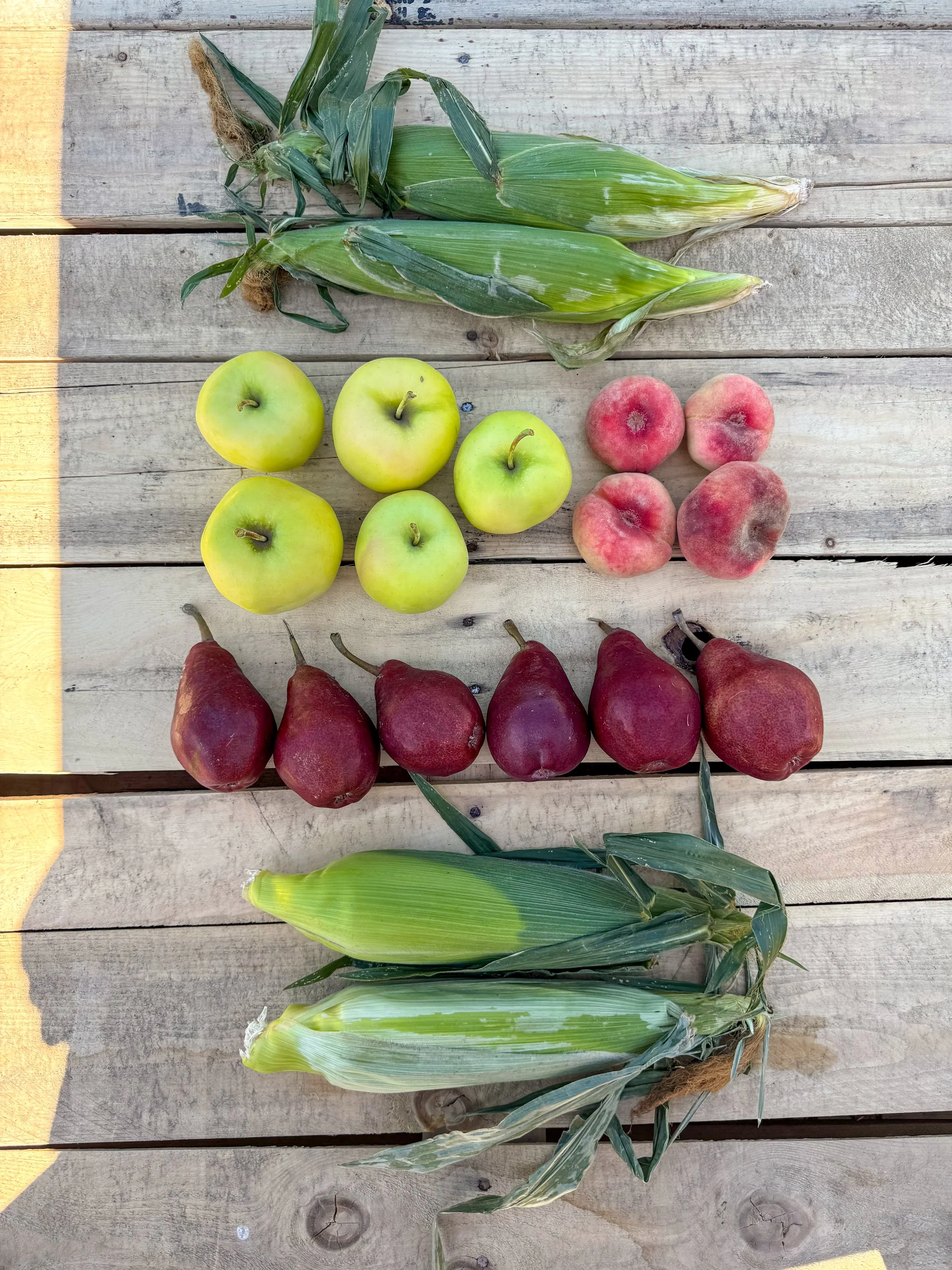 Six ears of corn, five green apples, four peaches, and six red pears are arranged on a light-colored wooden surface.