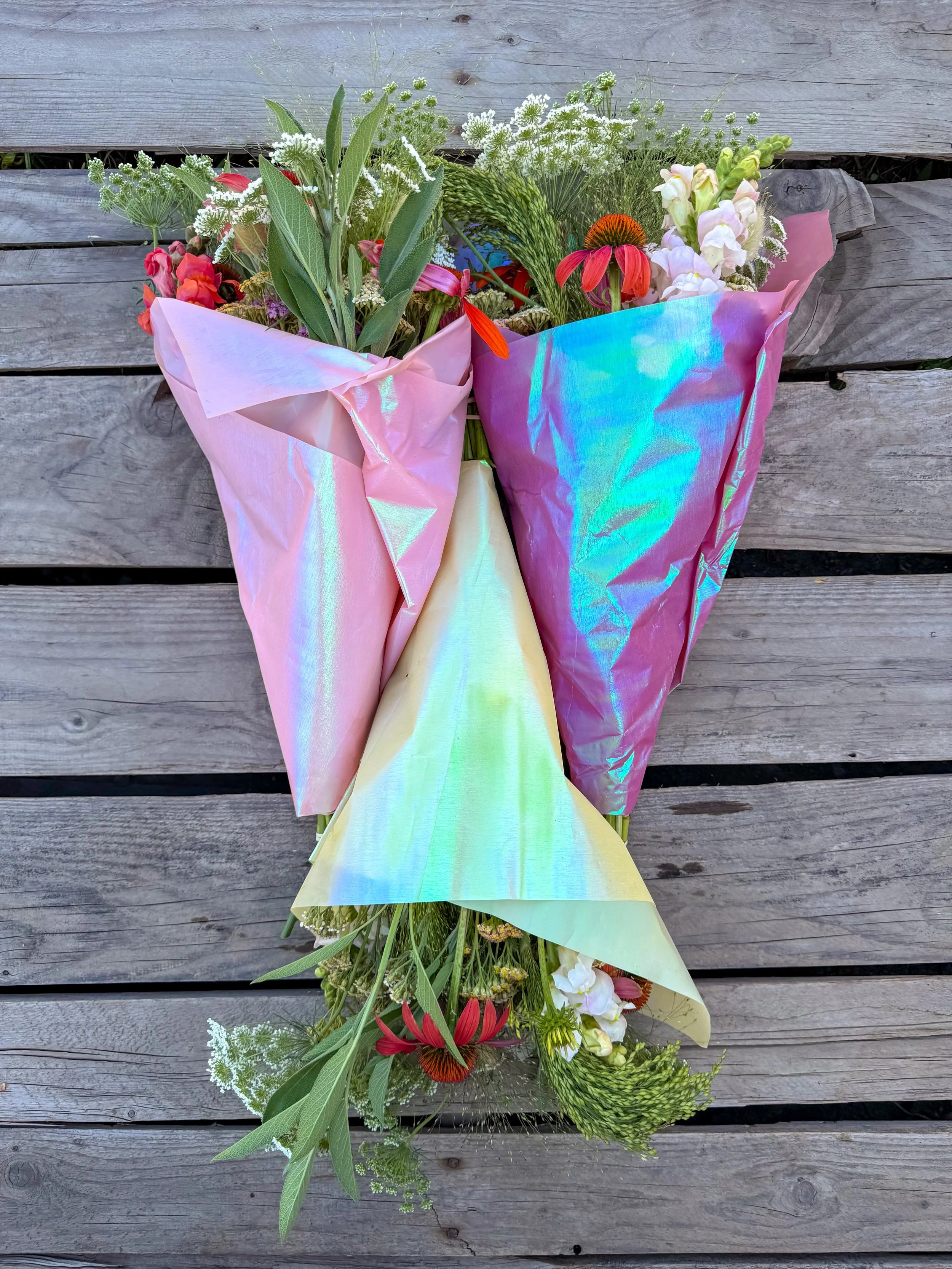 Three colorful bouquets of mixed flowers wrapped in pink, iridescent, and yellow wrapping paper, resting on a wooden surface.