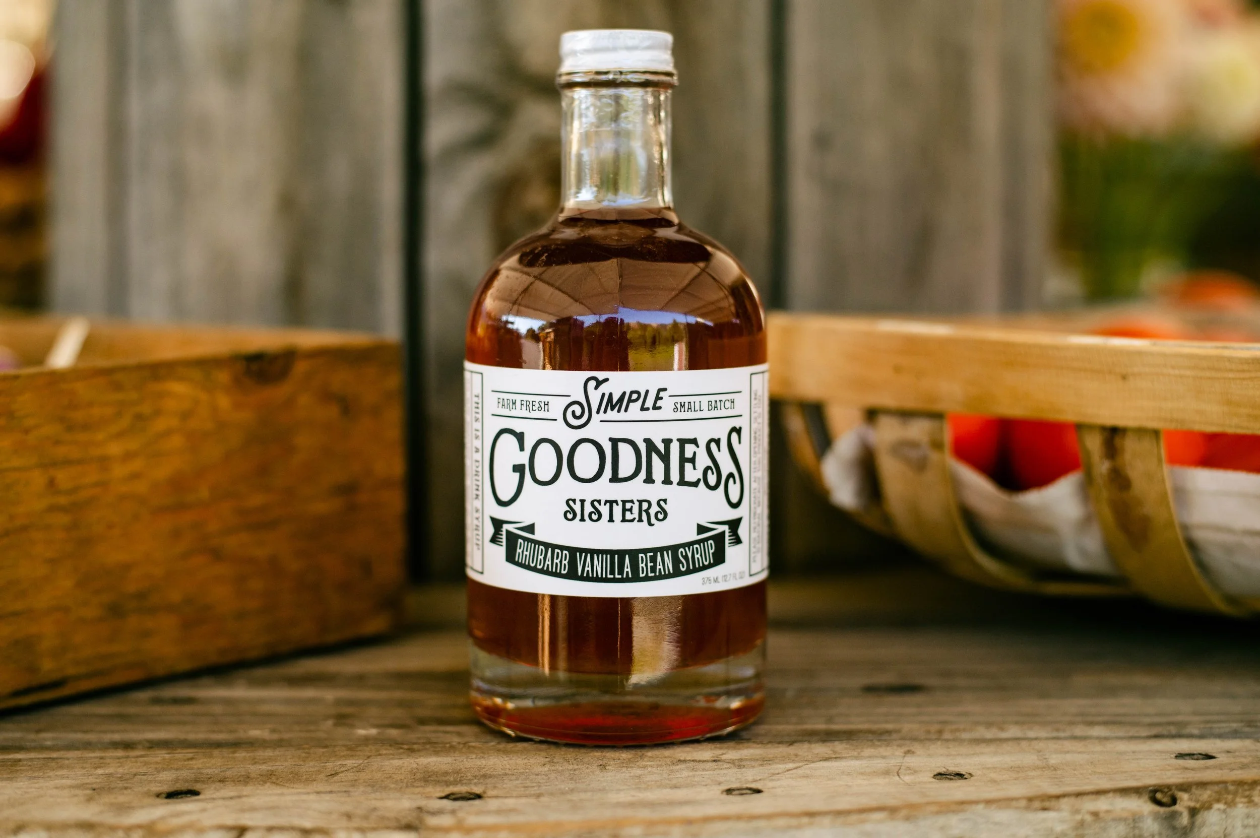 A glass bottle of Goodness Sisters Rhubarb Vanilla Bean Syrup on a rustic wooden surface, with a wooden crate and basket with red tomatoes in the background.
