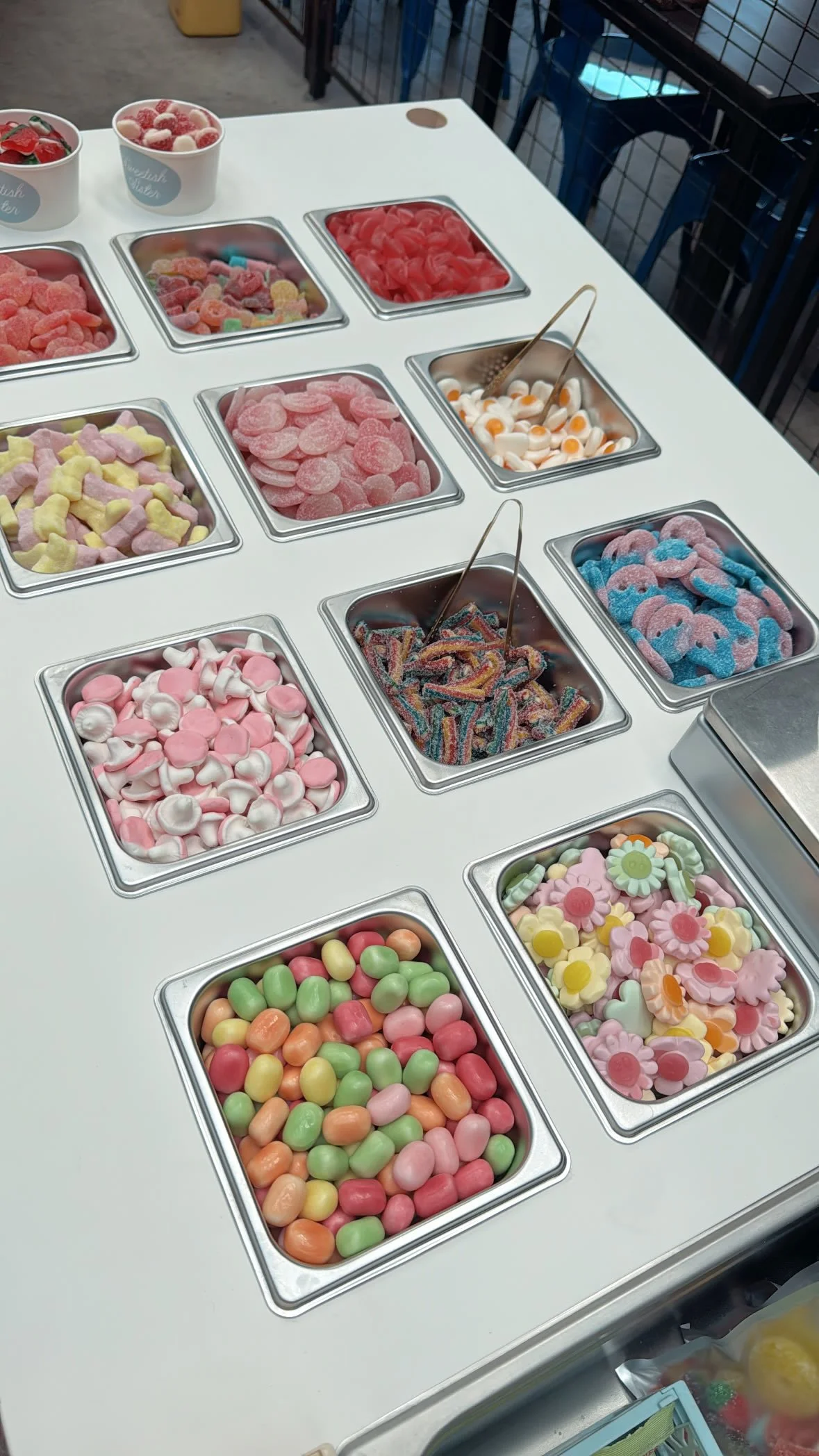 An assortment of colorful candies in stainless steel containers on a white table, including gummy worms, sour candies, marshmallow candies, and gummy bears.