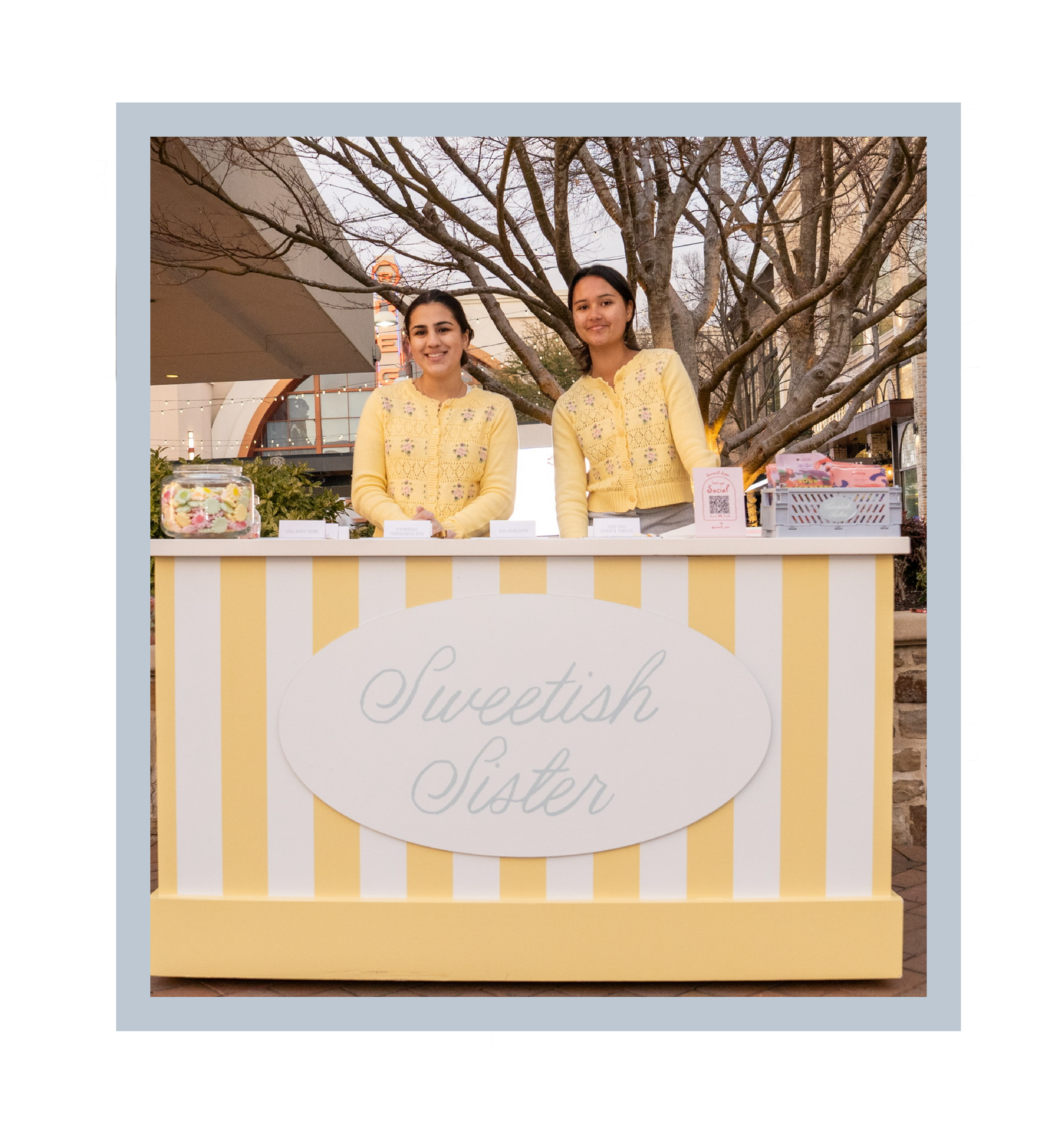 Two young women with dark hair wearing matching pastel yellow knitted cardigans standing behind a table with desserts at an outdoor market, under a large leafless tree during evening.