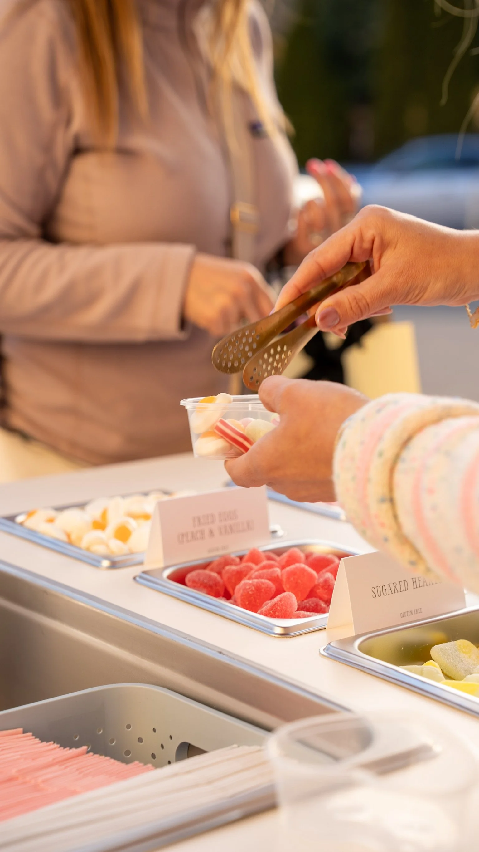 Close-up of a person serving gummy candies from a tray at a candy buffet. The candies are pink, yellow, and white. In the background, there is another person with blonde hair and a beige jacket, and various other candy trays with labels.