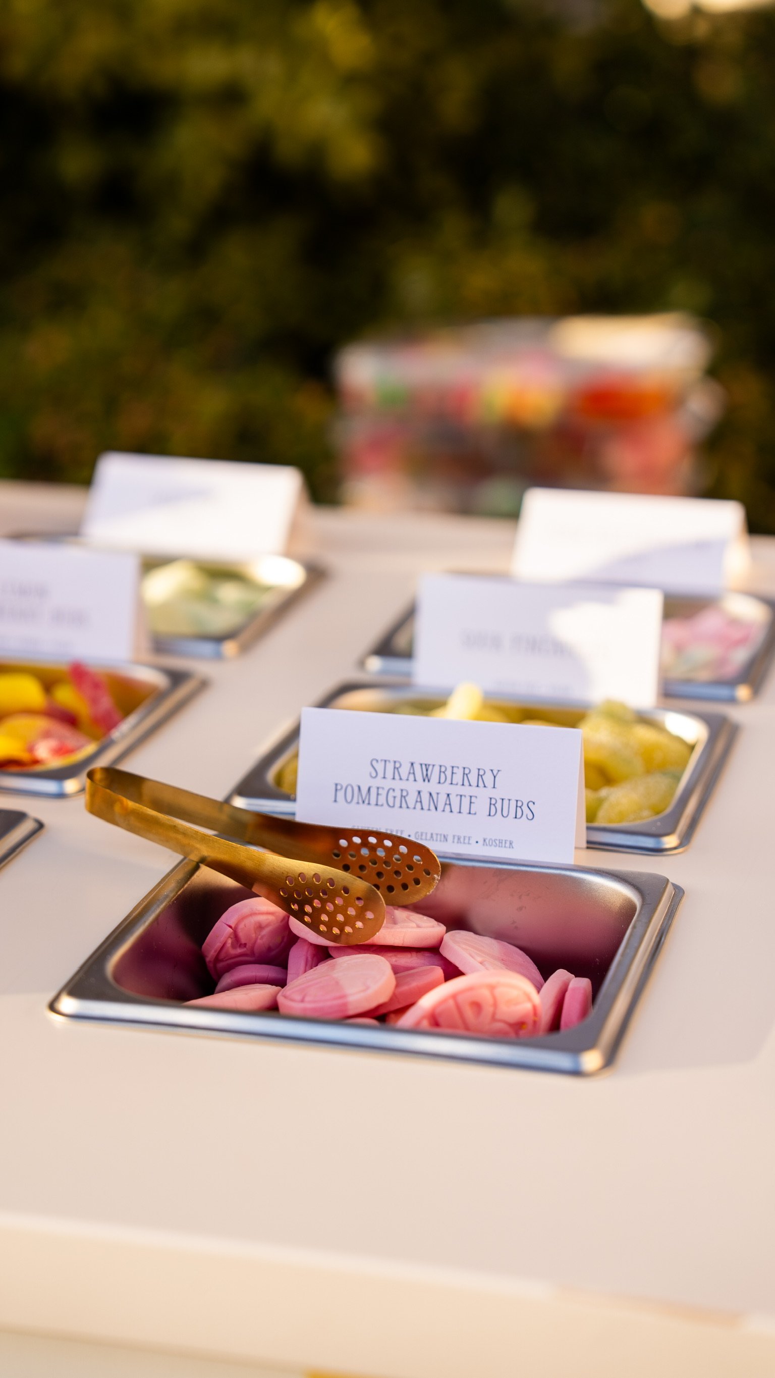 A buffet table with metal trays of sliced fruits, including strawberries, pomegranate buds, and pink onions, with labels indicating the fruit types.