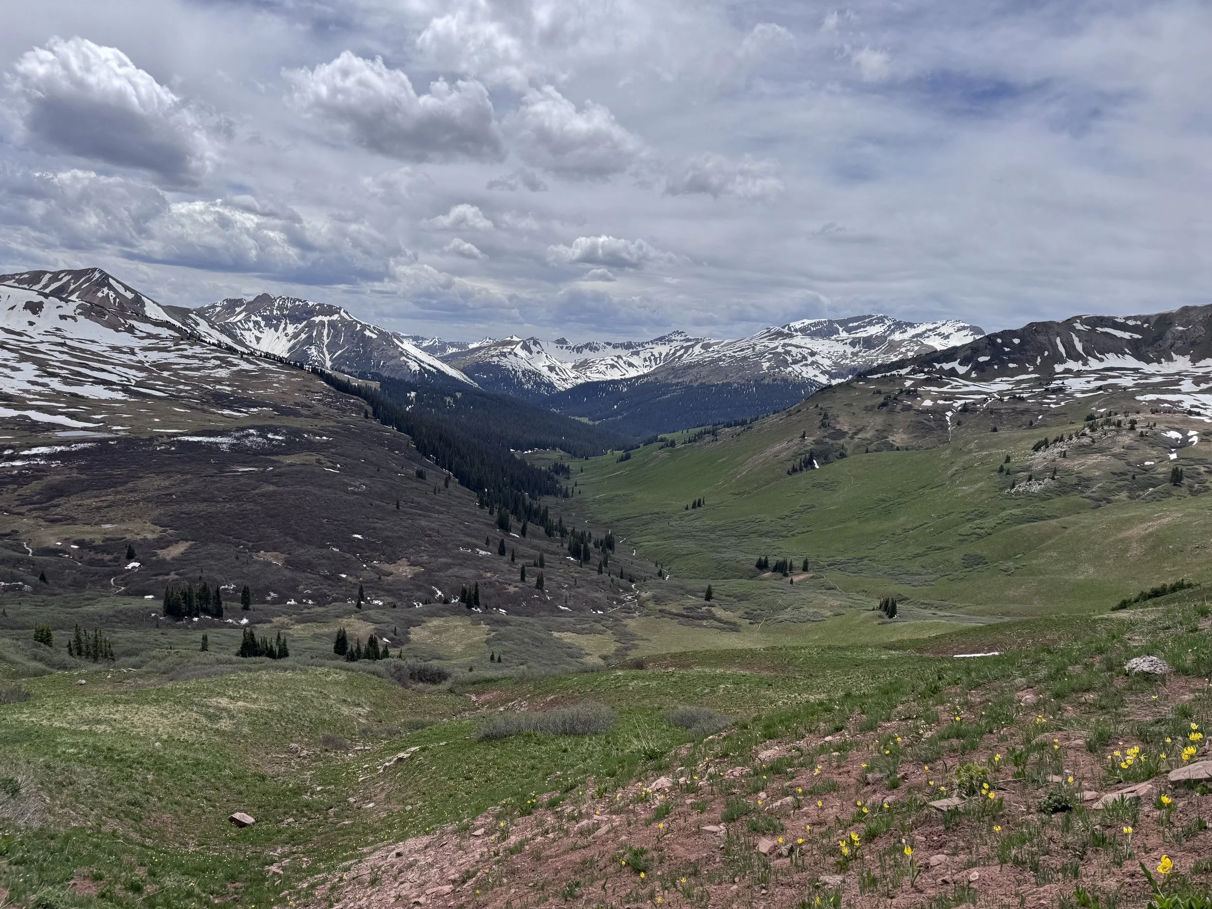 Mountain range with snow patches, green valleys, and cloudy sky.
