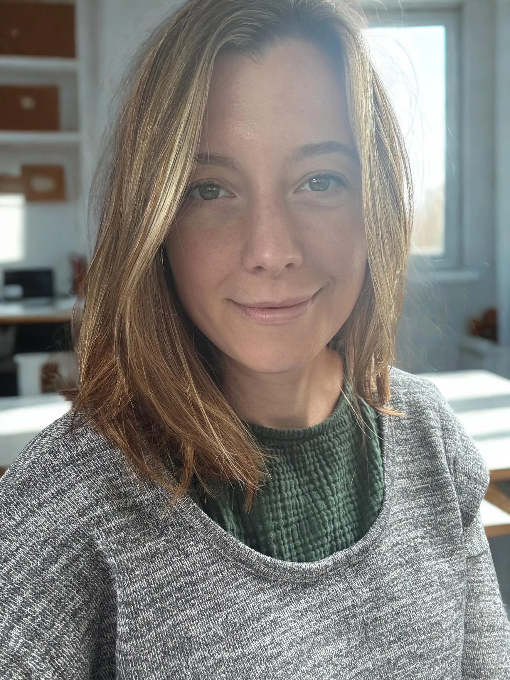 Close-up of a woman with long, light brown hair, blue eyes, and a friendly expression, wearing a gray and green sweater, inside a well-lit room with a window and bookshelf in the background.