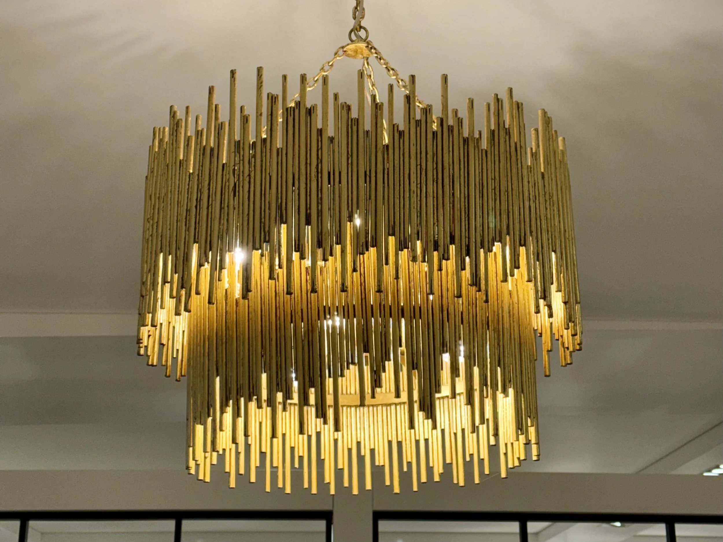 Large gold chandelier with numerous hanging gold rods, illuminated from within, hanging from a chain attached to a ceiling.