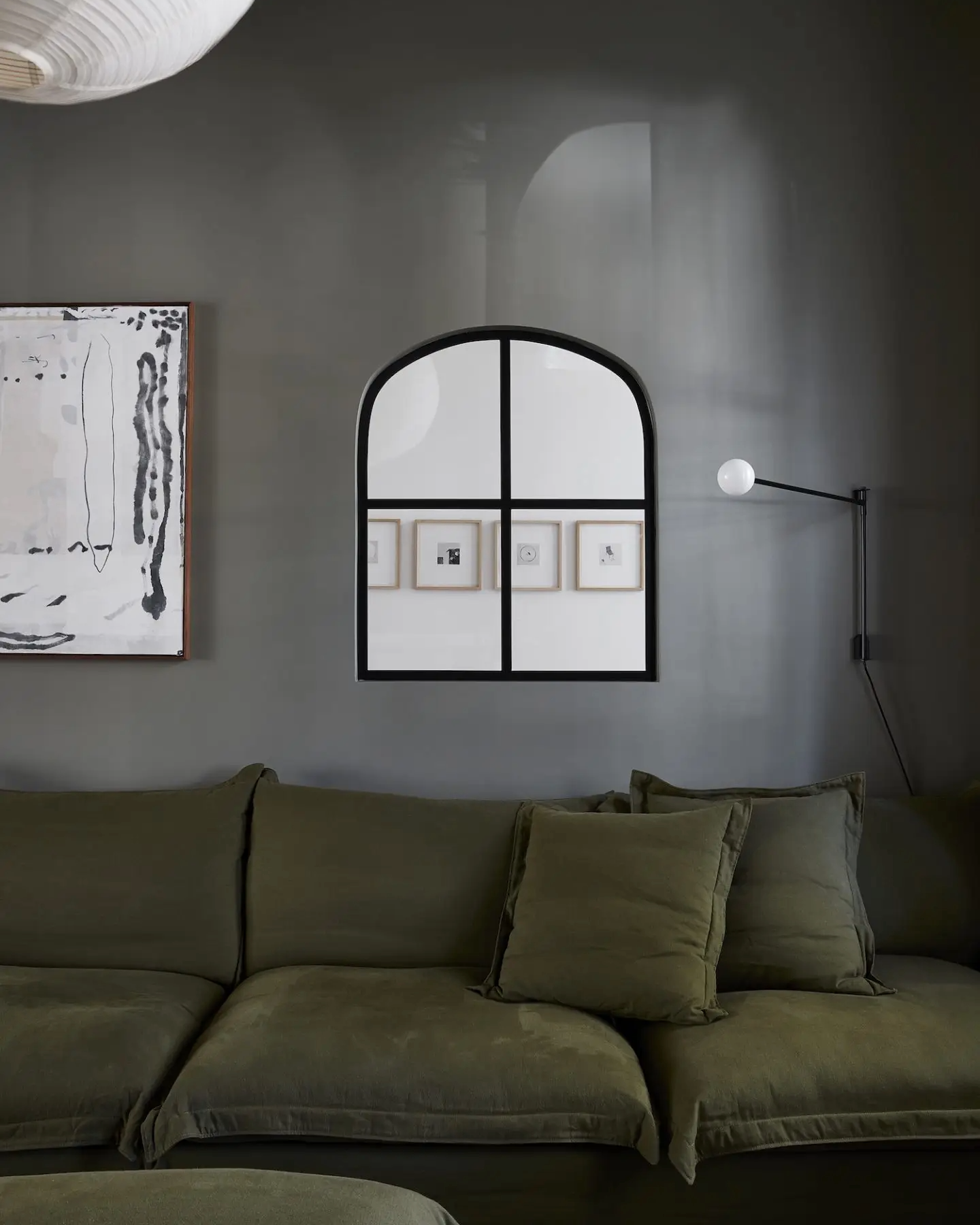 A modern living room with a green sofa, abstract artwork on the wall, black-framed arched window, and a wall-mounted black lamp with a white globe bulb.