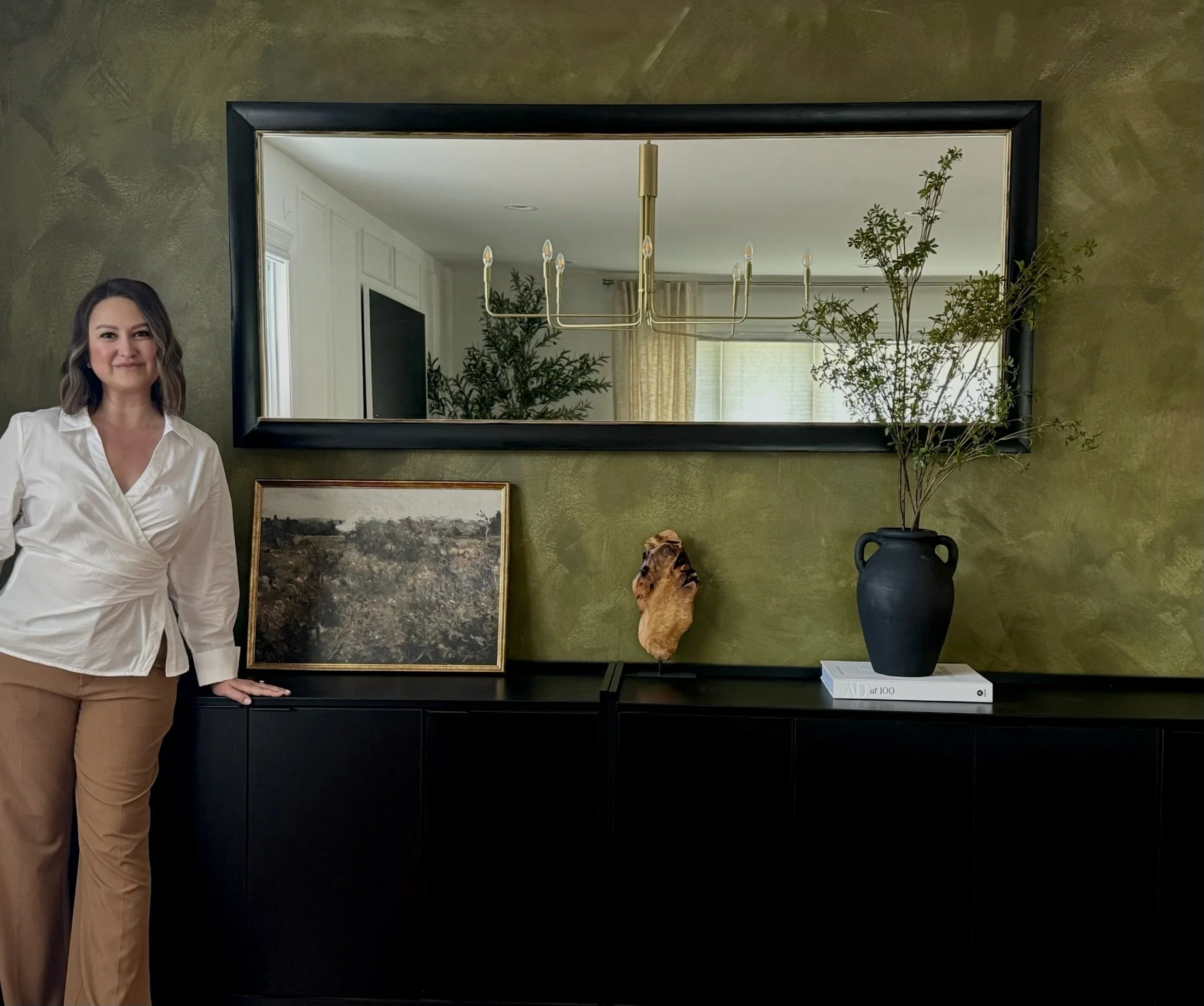 Martina Kloss, a woman in a white blouse and tan pants standing next to a black sideboard with decorative items, a large mirror above, a framed landscape painting, a carved wooden sculpture, and a black vase with branches on top.
