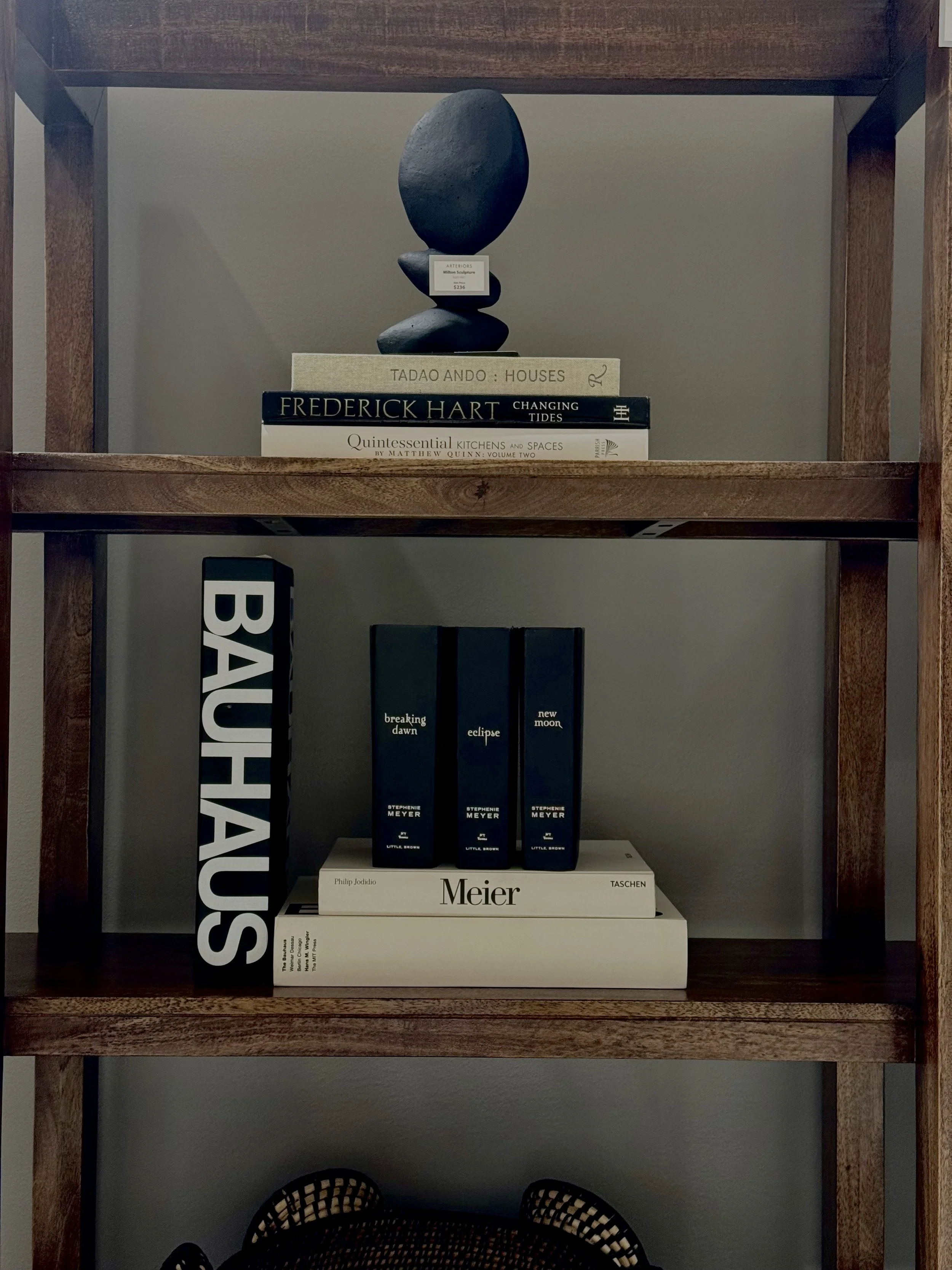 A wooden bookshelf with two shelves holding books and decorative items. The top shelf has a stack of books and a stacked stone sculpture on top. The bottom shelf features books standing upright with a large black and white book sign saying 'BAUHAUS' and a trio of Stephenie Meyer books titled 'Breaking Dawn,' 'Eclipse,' and 'New Moon.'
