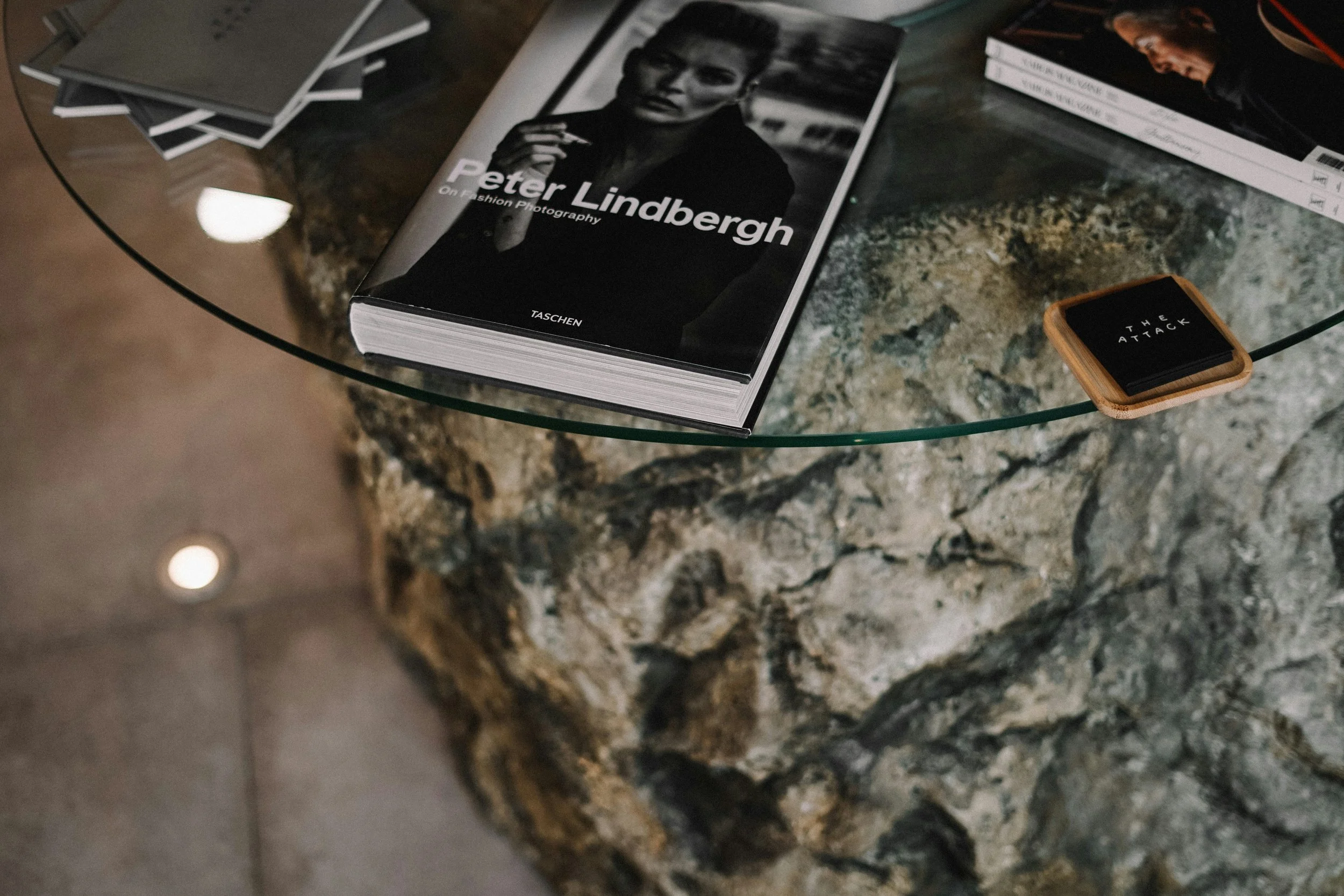A glass-top table with a thick, irregularly shaped stone or concrete base. On the table are several coffee table books, including one about Peter Lindbergh, and a small stack of magazines or brochures on the left. There is also a black coaster with white text on a wooden tray or coaster holder.