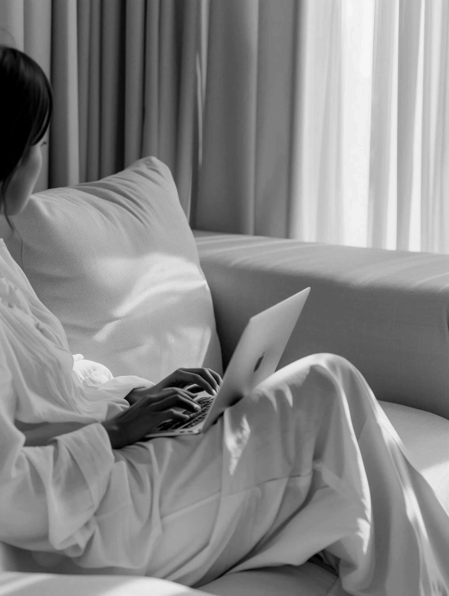 A woman sitting on a sofa using a laptop, dressed in white clothing, with curtains in the background.