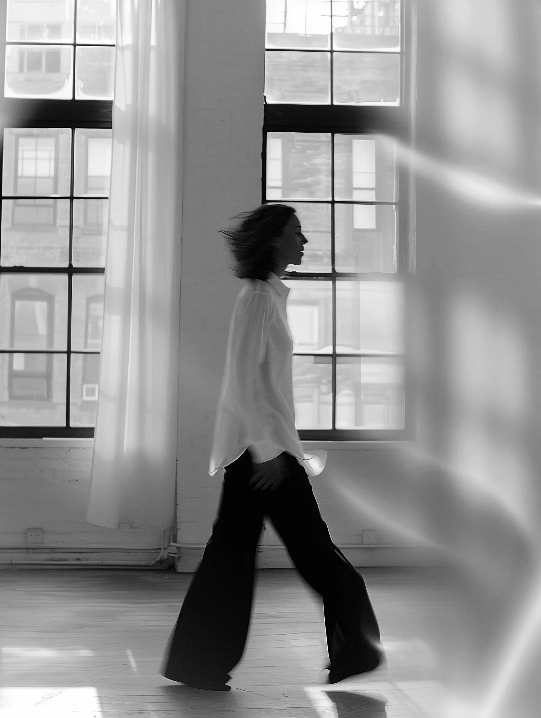 A woman walking indoors in front of large windows with sunlight streaming in, captured in black and white.