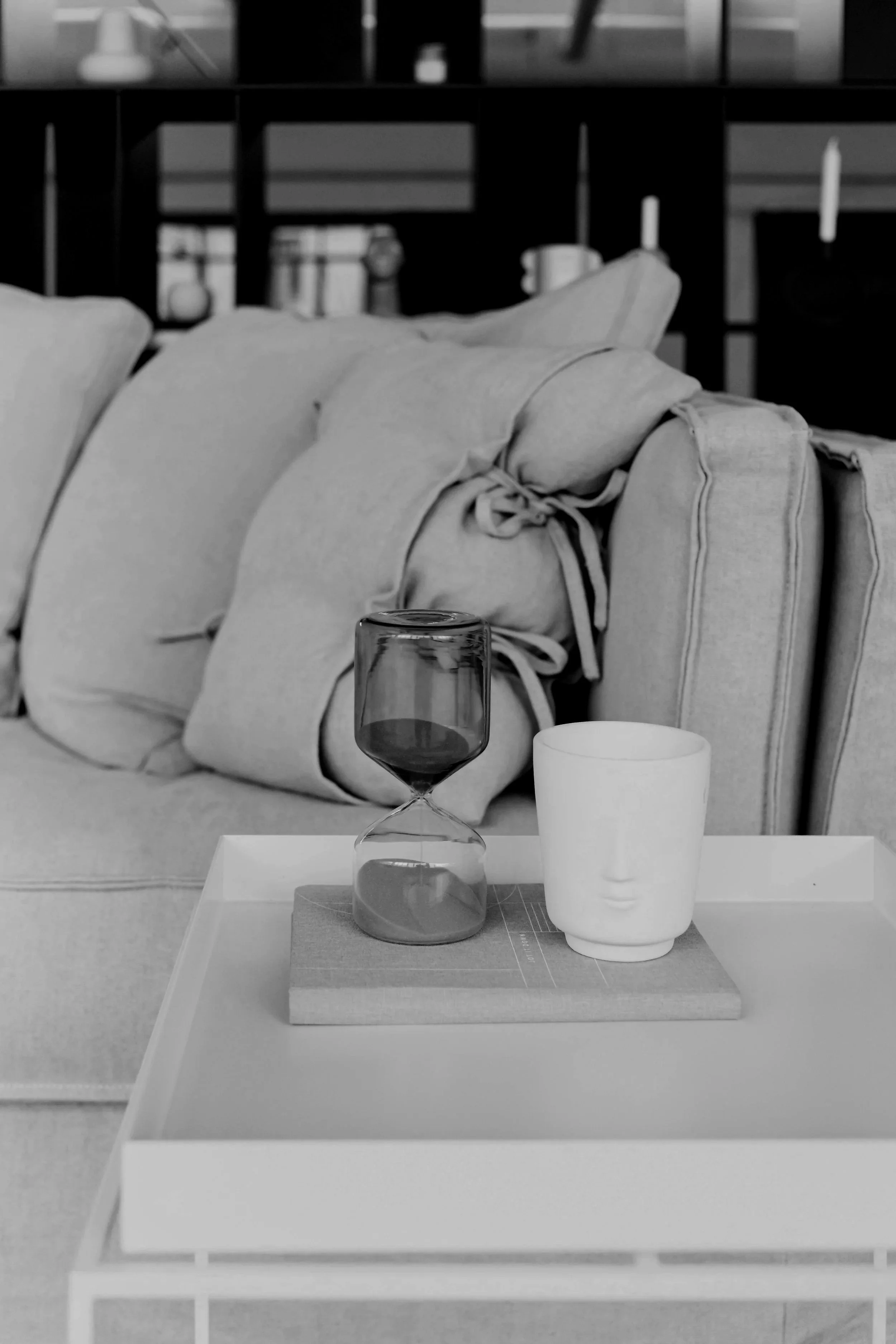A white side table with a book, an hourglass with dark sand, and a white ceramic cup with a face design. In the background, a sofa with pillows and a bookshelf with decorative items are visible.