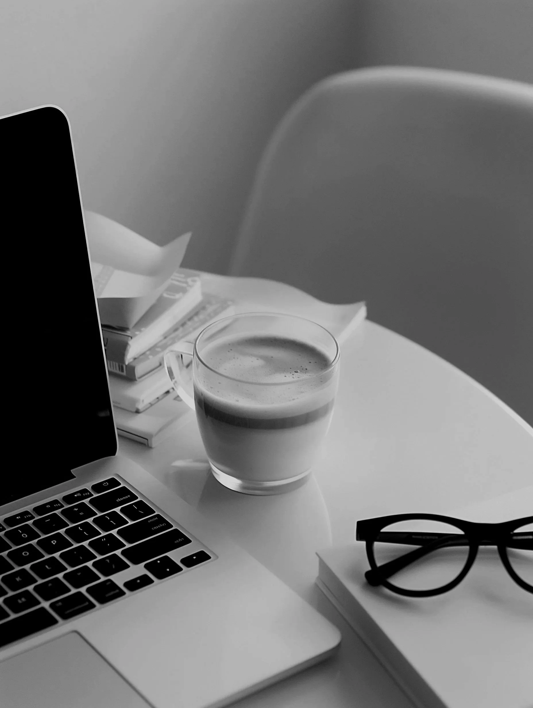 Black and white photo of a laptop, a cup of coffee, glasses, and books on a white table.