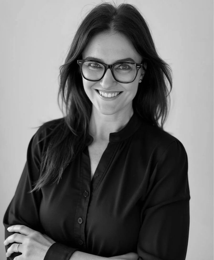 Black and white portrait of a woman with dark hair, wearing glasses and a black shirt, smiling with arms crossed.