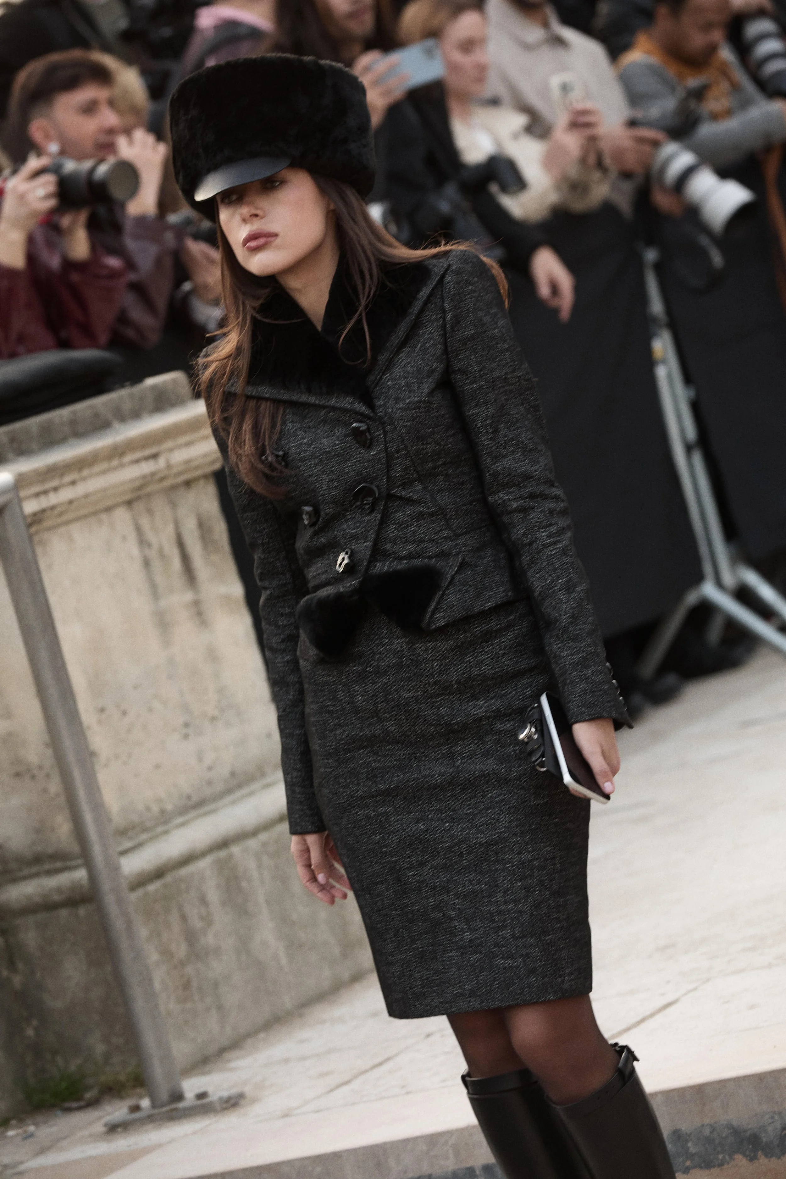 A woman wearing a dark gray coat, matching skirt, black knee-high boots, and a large black fur hat stands outdoors on a city street, holding a smartphone in her right hand, with people and photographers in the background.