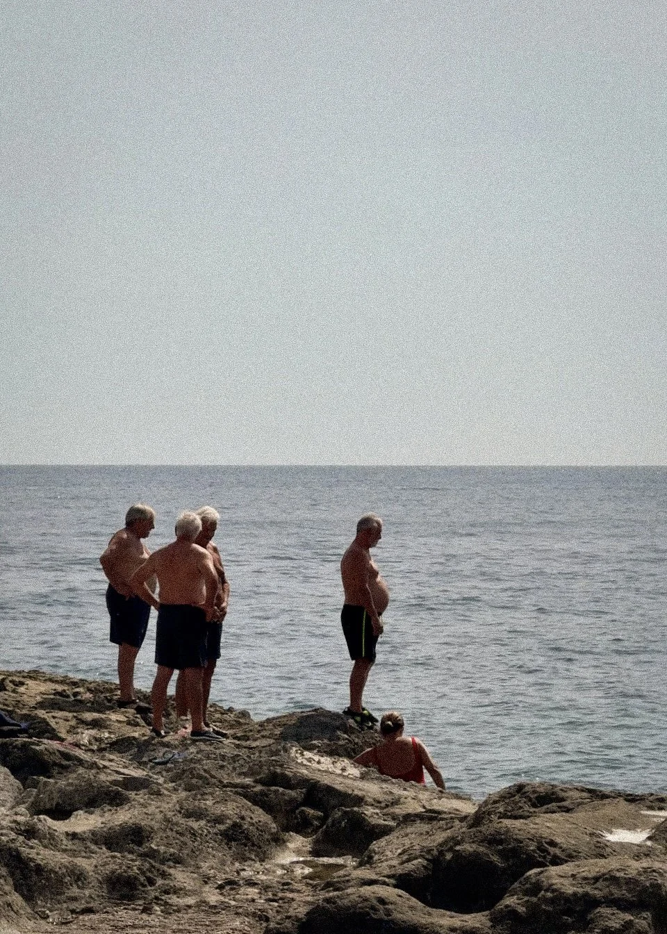A group of elderly Italians standing and sitting on rocks by the ocean in Puglia, Italy, looking out at the water on a sunny day.