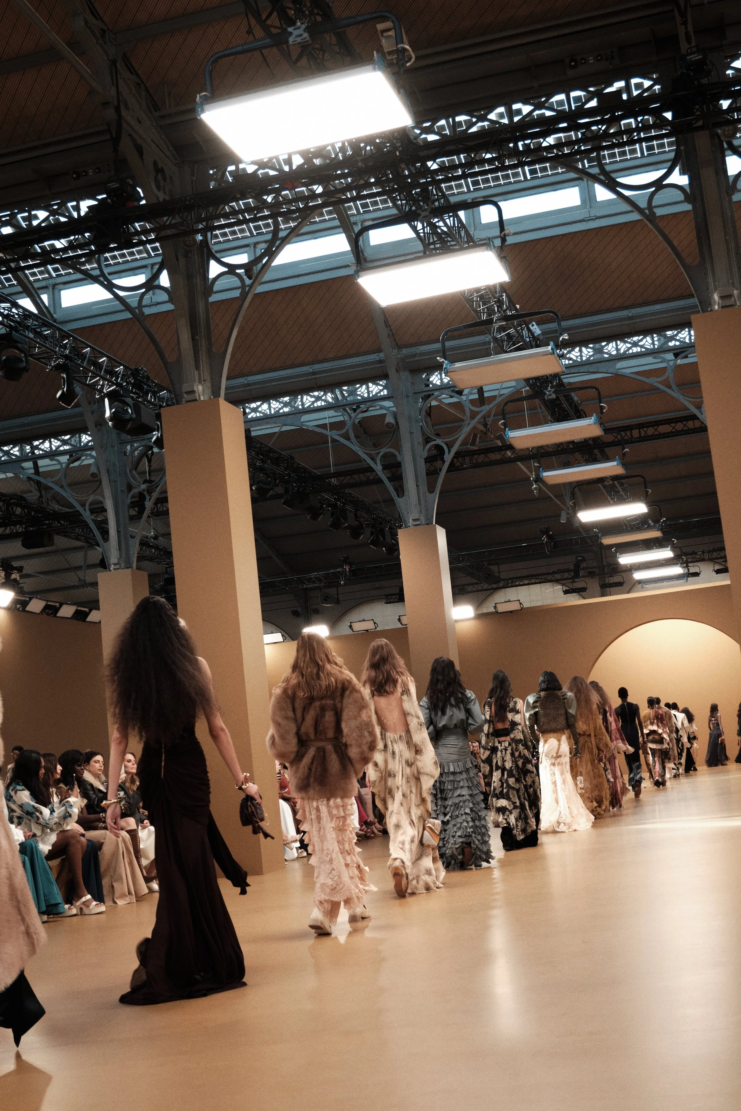 Models walking down runway during a fashion show in a large, industrial-style venue with high ceilings, metal beams, and overhead lighting.