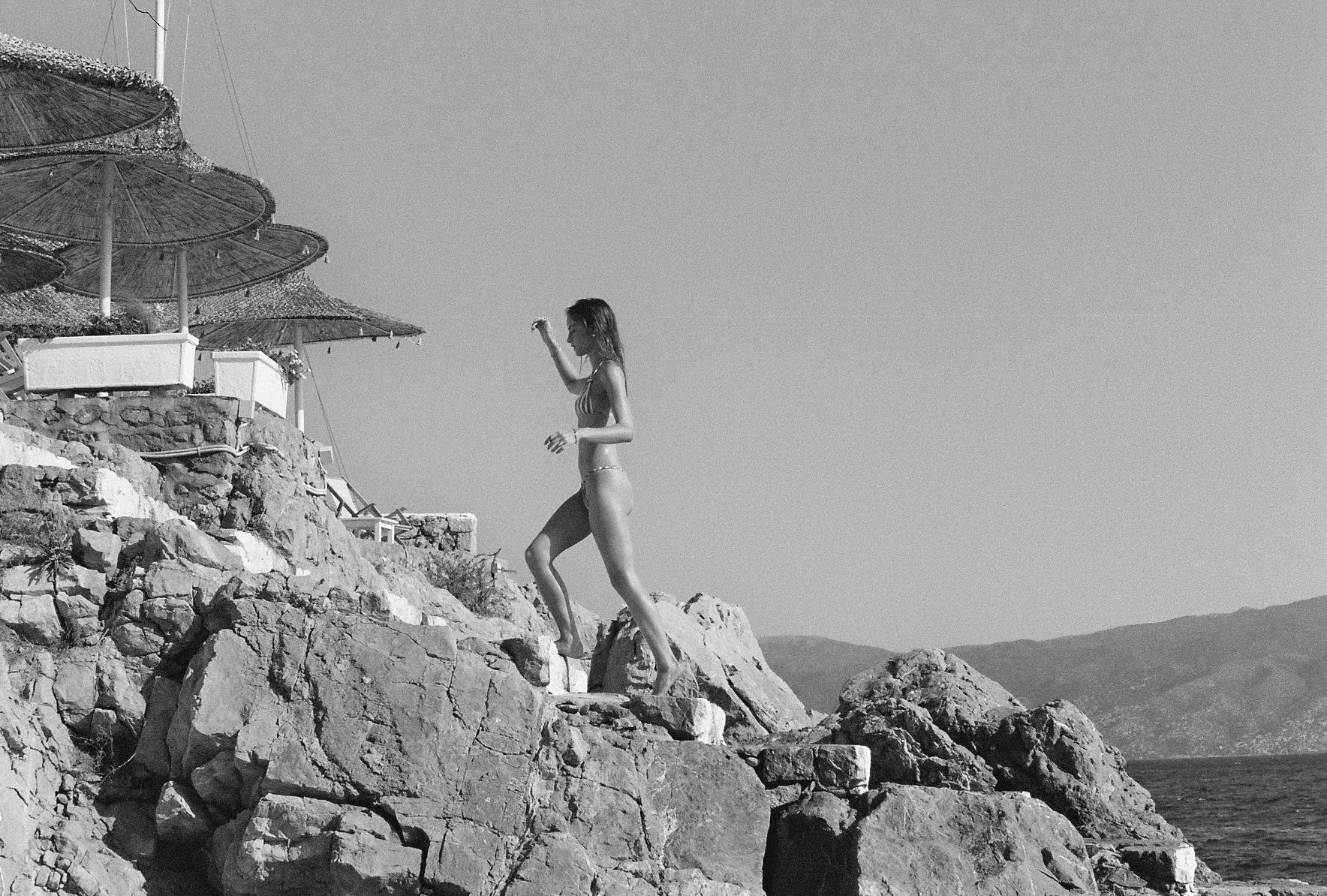 A woman in a bikini walking on rocky terrain by the seaside with umbrellas and chairs in the background.