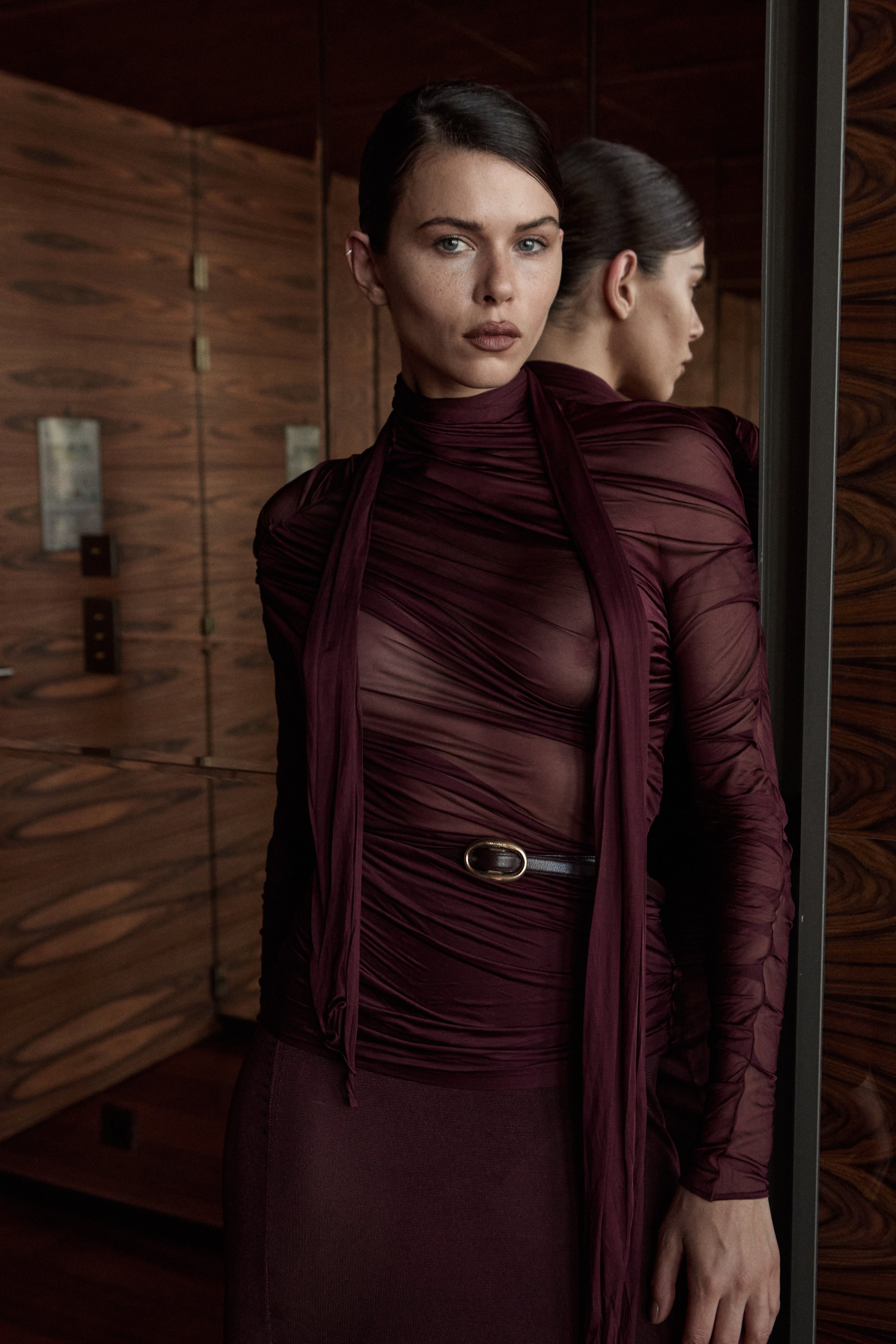 Model Georgia Fowler standing in front of a mirror, wearing a sheer maroon top with ruched fabric and a black belt, with wooden paneled walls in the background.