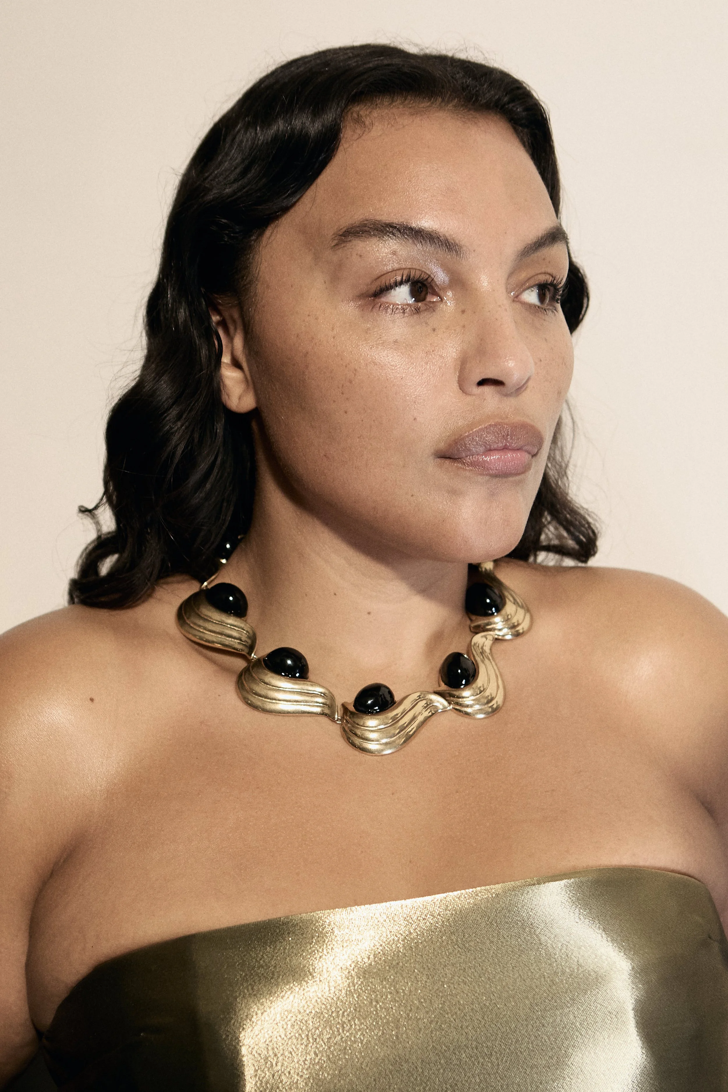 A woman with dark hair wearing a strapless metallic dress and a gold necklace with black accents.