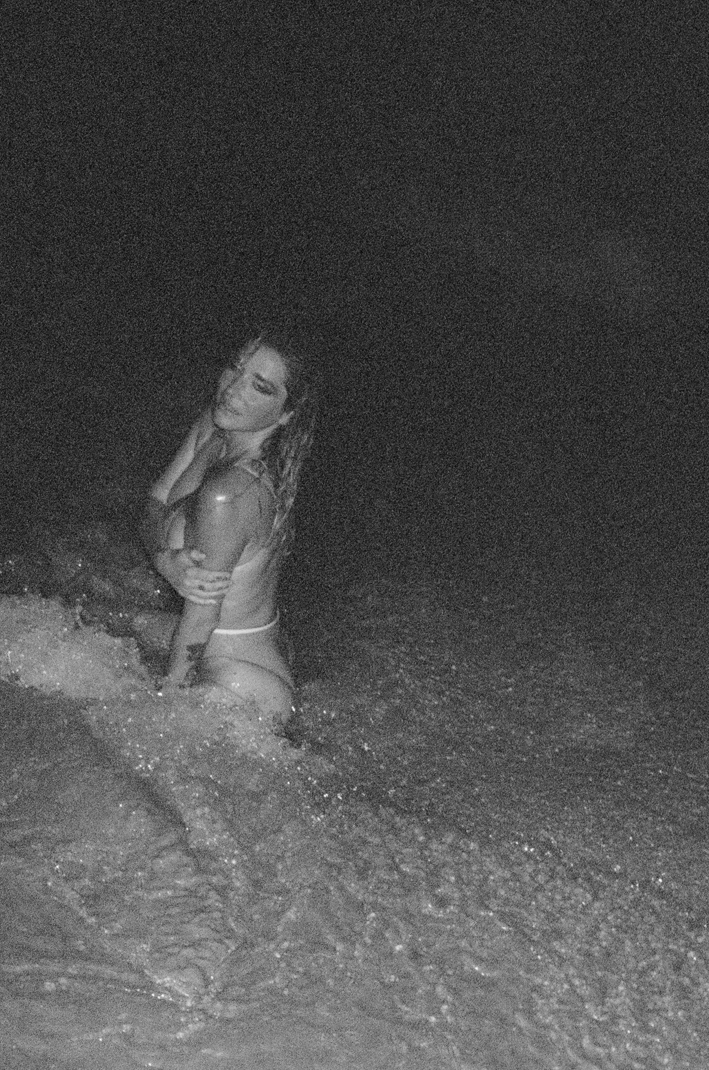A woman in a swimsuit sitting in shallow water at night, with her eyes closed and head tilted back.
