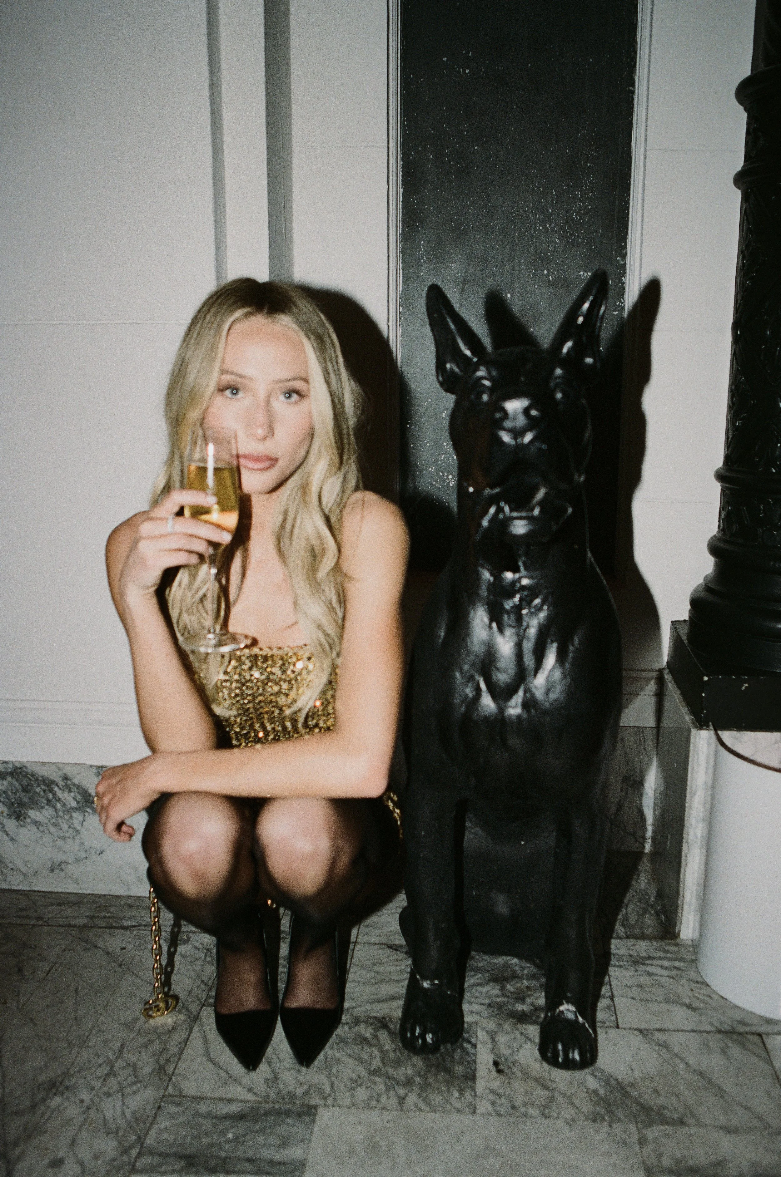 A woman with long blonde hair wearing a gold sequin dress and black high heels, holding a glass of champagne, squatting next to a black statue of a dog in an indoor setting.