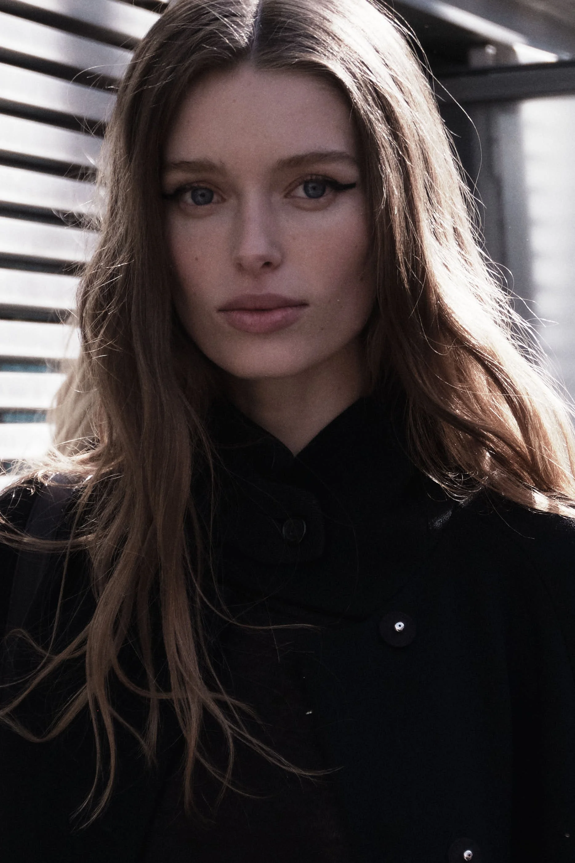 Close-up of a young woman with long wavy brown hair, blue eyes, and makeup, wearing a black shirt, outdoors with sunlight on her face.