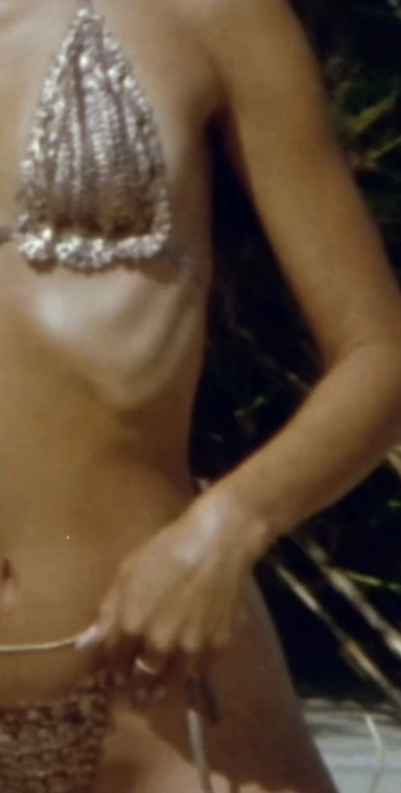 Close-up of a person's torso with a silver, bejeweled bra and a hand holding a thin stick or rod.