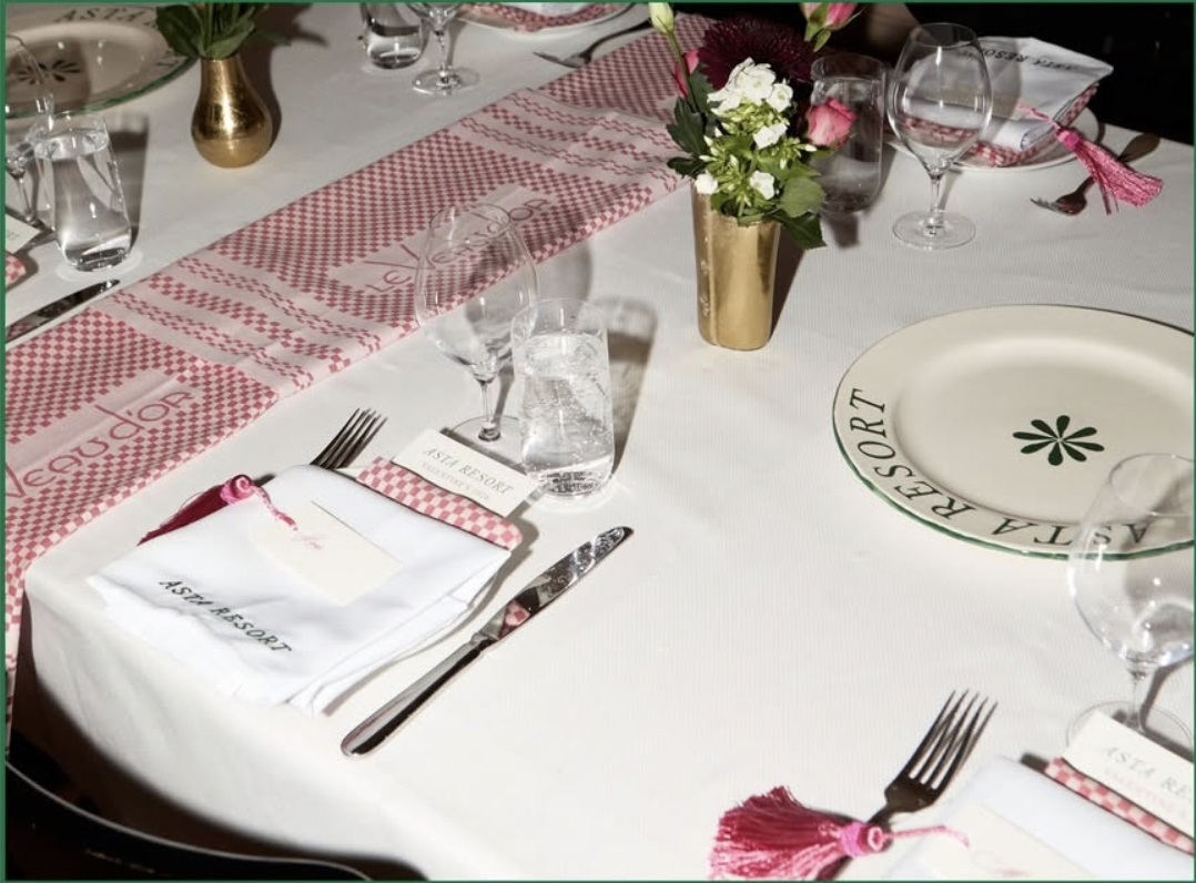 Formal dining table setup with white tablecloth, pink checked table runner, gold vase with flowers, glassware, silverware, and place settings.