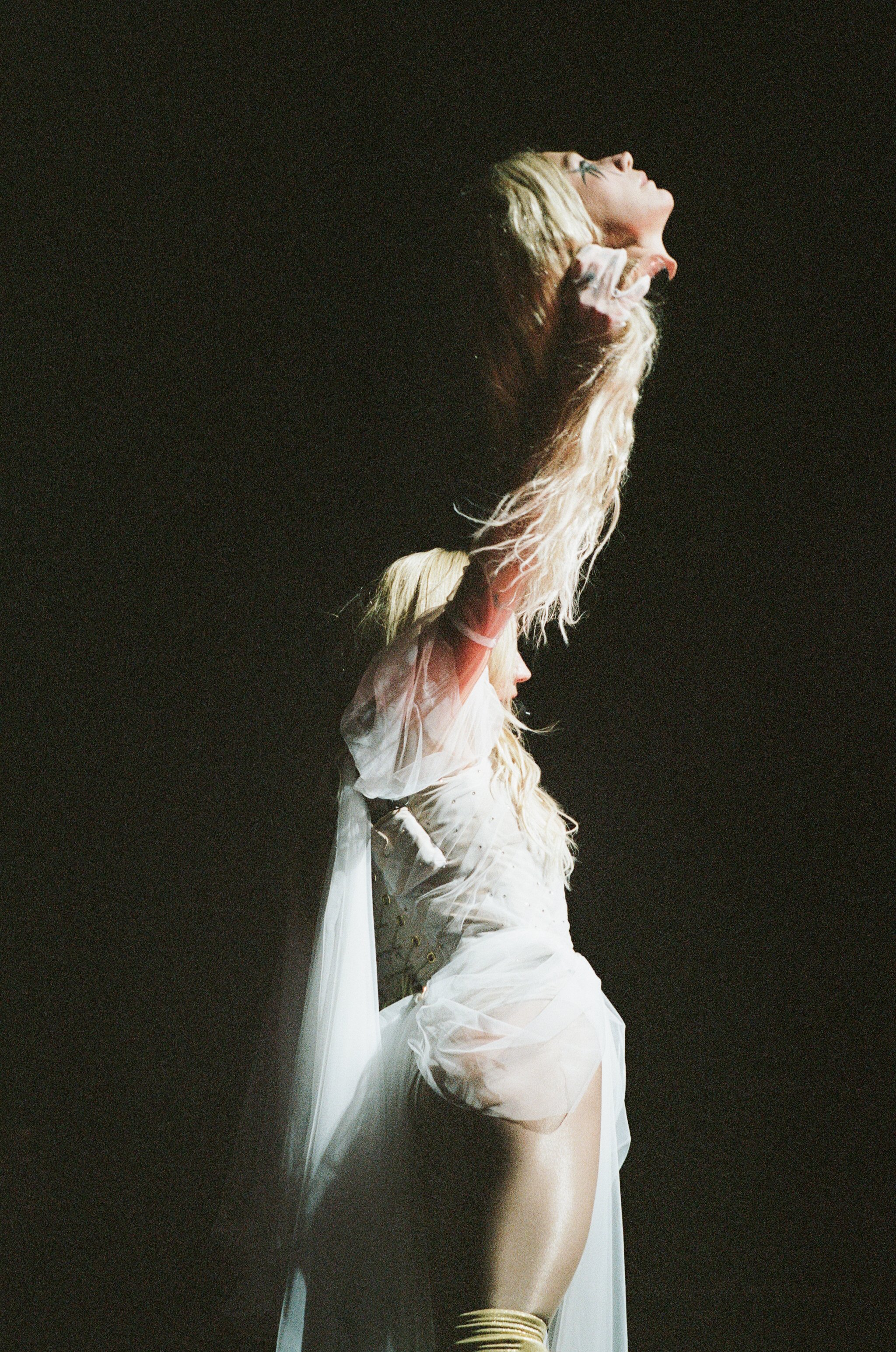 A woman with blonde hair is performing on stage, holding a microphone in her right hand, with her head tilted back and eyes closed, dressed in a sheer white outfit with puffy sleeves and gold accessories.