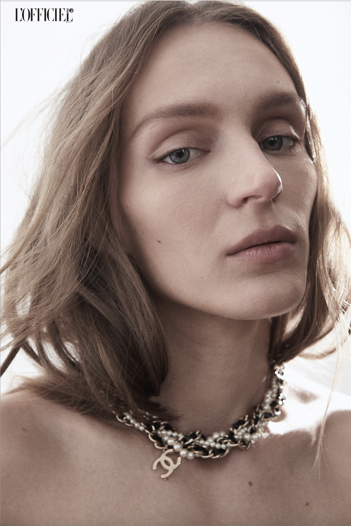 Close-up of a young woman with shoulder-length brown hair, wearing a Chanel necklace, with a neutral expression, for L'Officiel magazine.