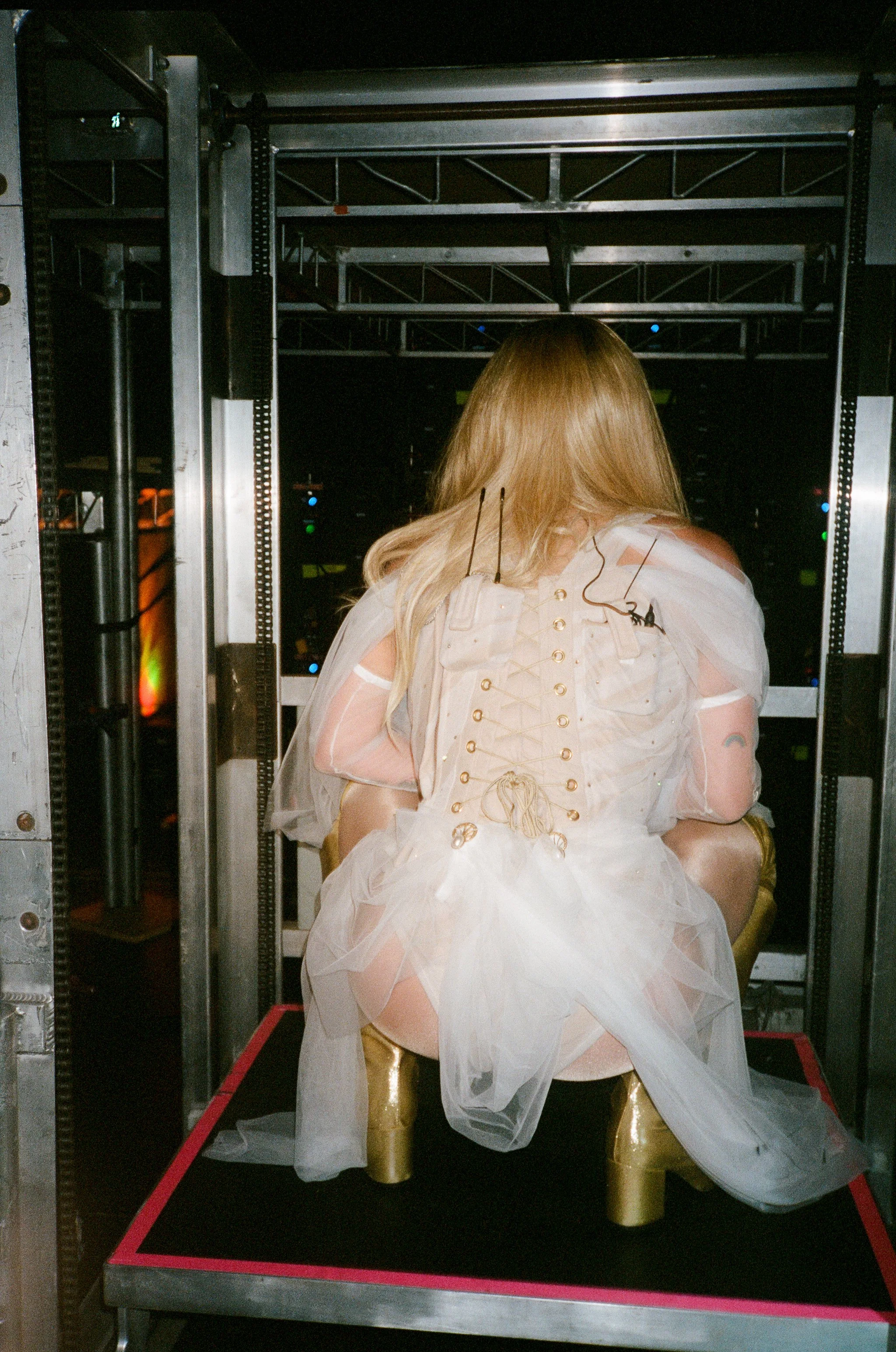 A person with long blonde hair, dressed in a white, sheer, laced costume, seated on a gold chair in front of a server rack, with their back facing the camera.