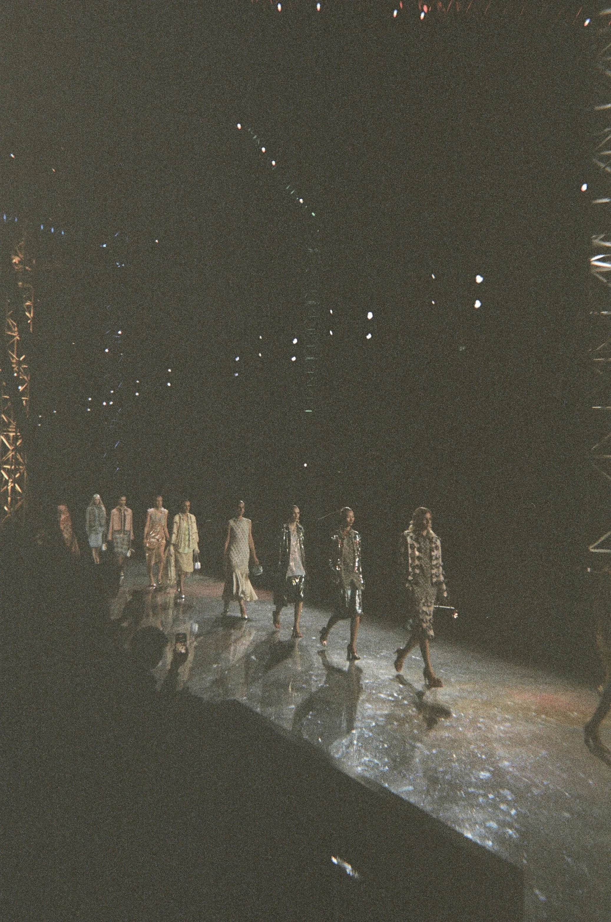 Models walking down a reflective runway at a fashion show, under dark lighting with spotlights.