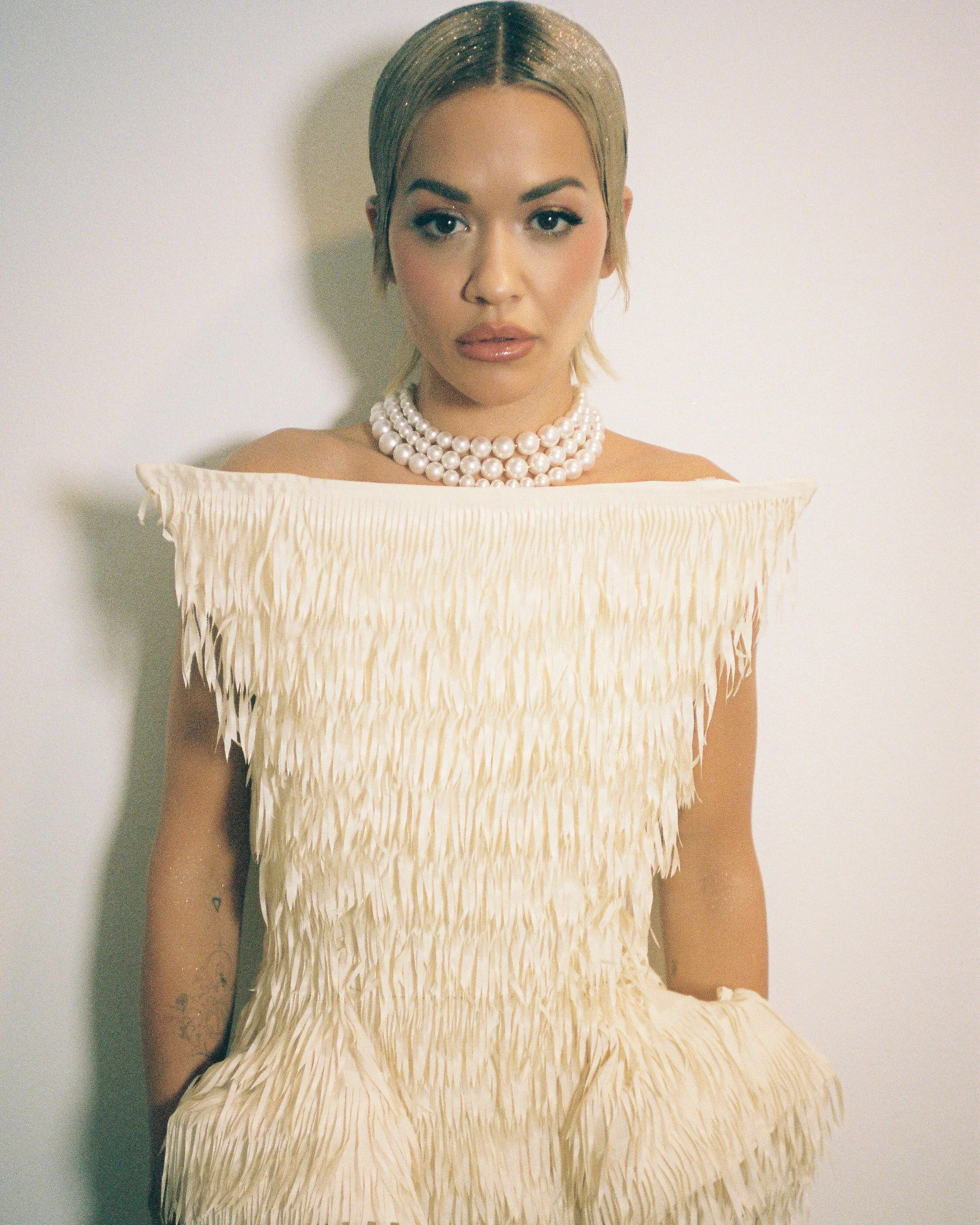 Singer Rita Ora A wearing a double strand pearl necklace and a cream-colored dress with textured feather-like details, standing against a plain white wall.